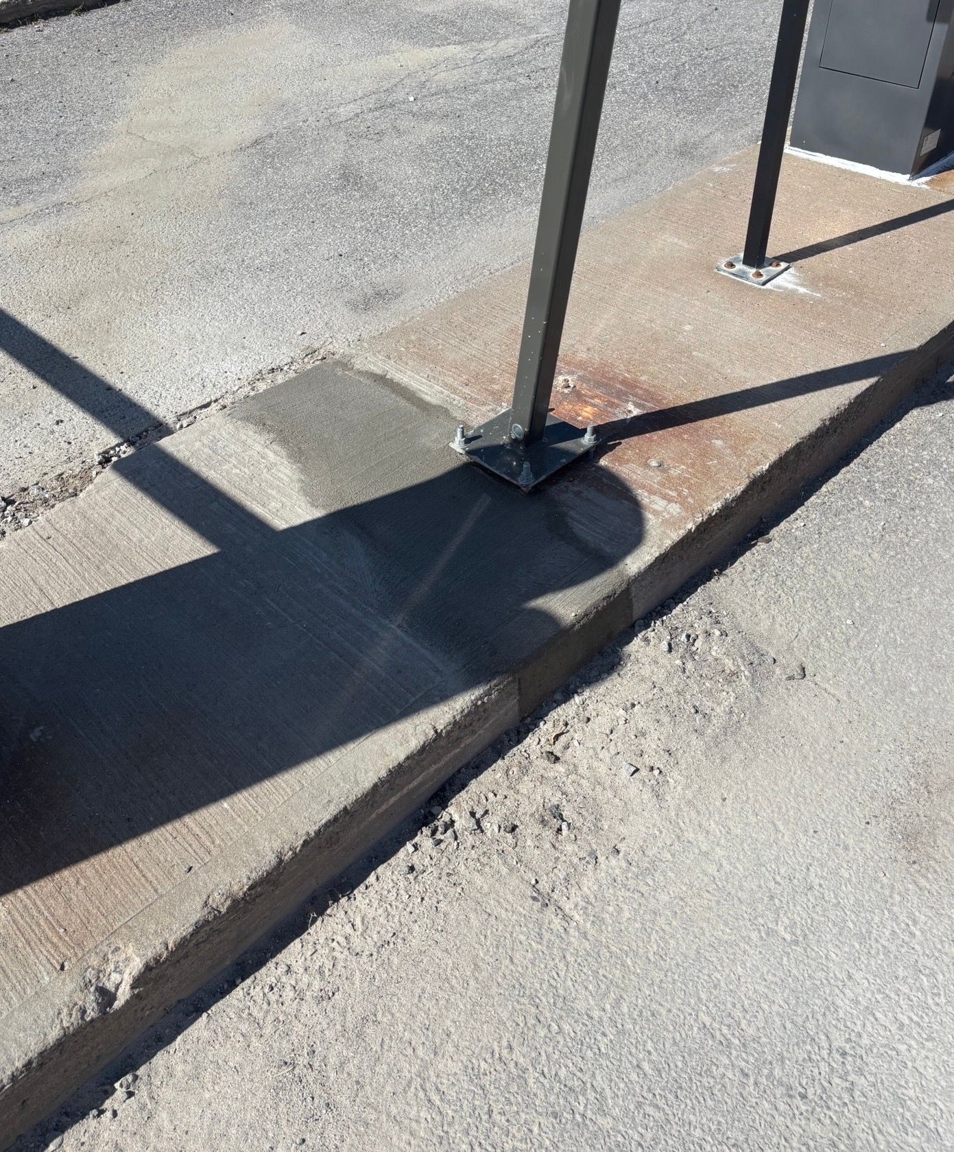 Trottoir en béton endommagé avec bordure fissurée et socle de poteau métallique projetant une ombre
