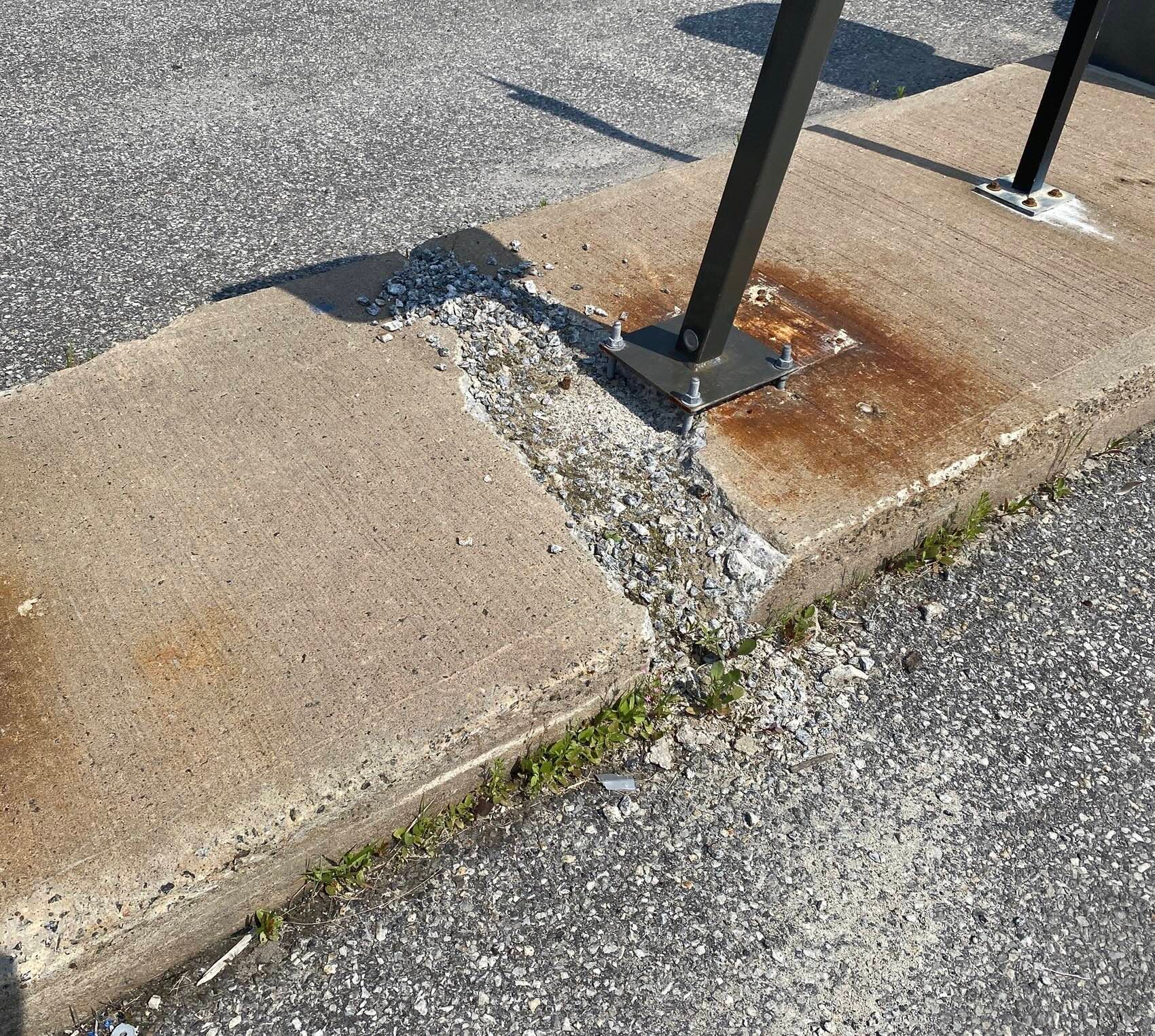 Bordure et trottoir endommagés, béton fissuré autour d'un socle de poteau métallique rouillé