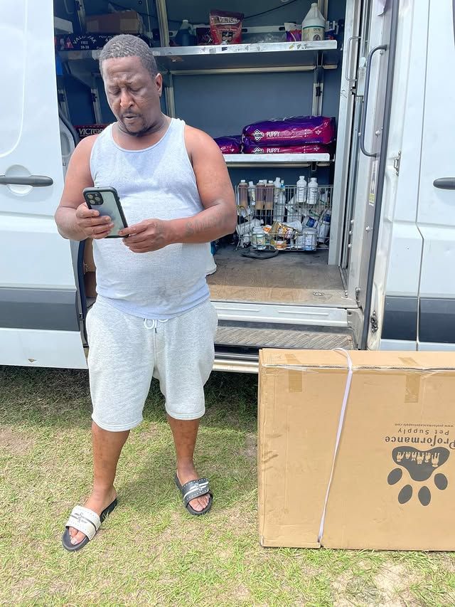 Man standing by an open van, looking at a phone, with boxes and supplies inside and beside the vehicle.