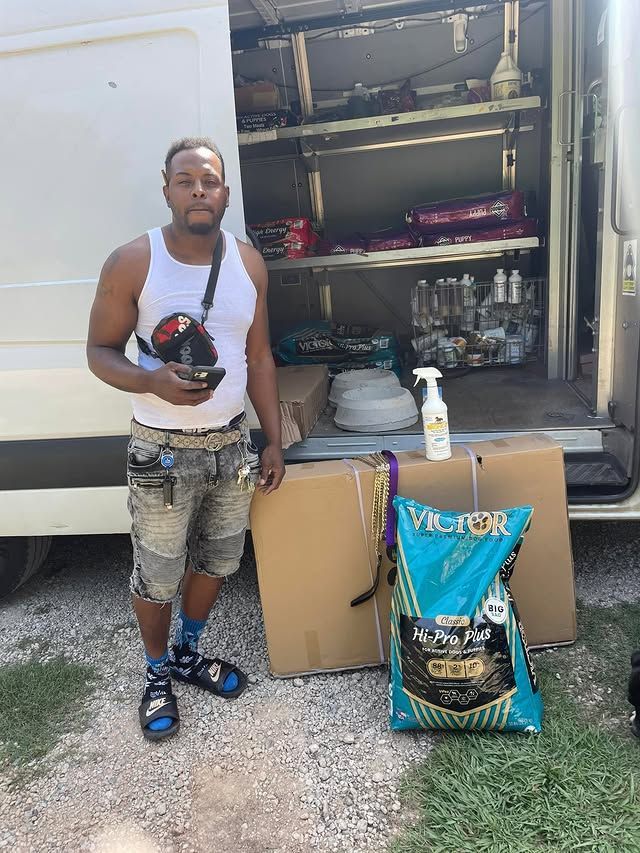 Man standing by a van loaded with supplies, holding a bottle, beside stacked boxes and a large pet-food bag