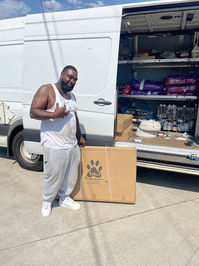 Man standing beside a white cargo van, holding a cardboard box in bright daylight.
