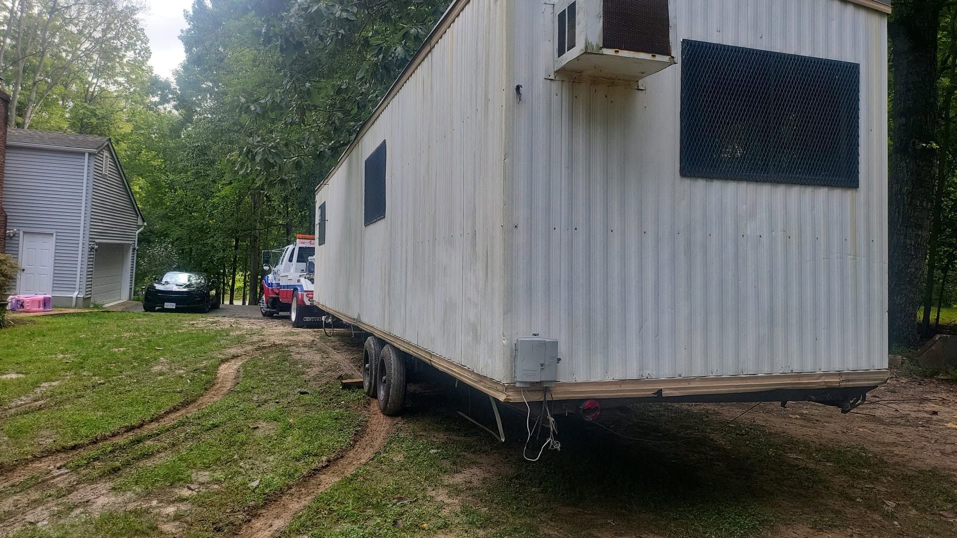 White mobile trailer on a grassy dirt slope, with a tree-lined yard and a house in the background.