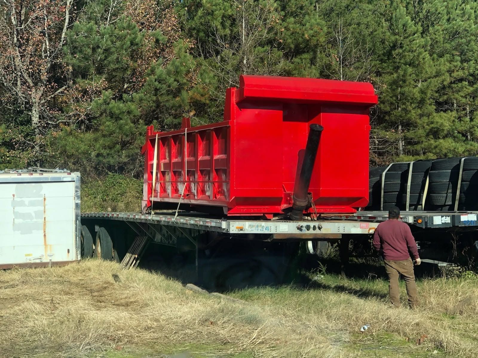 Red dump trailer on a flatbed truck in a grassy field, with a person standing nearby.