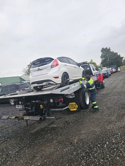 White SUV being loaded onto a tow truck in a gravel lot on a rainy day, with workers nearby
