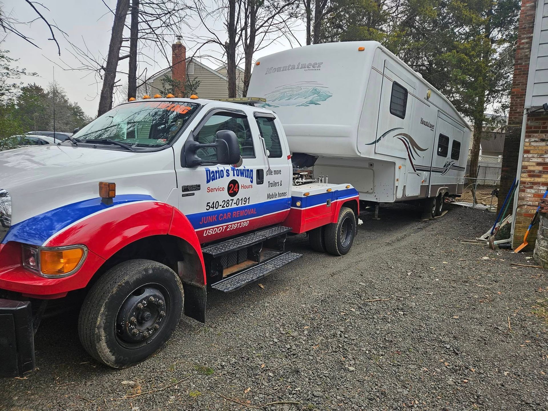 Tow truck towing a white RV on a gravel roadside near trees and a utility pole