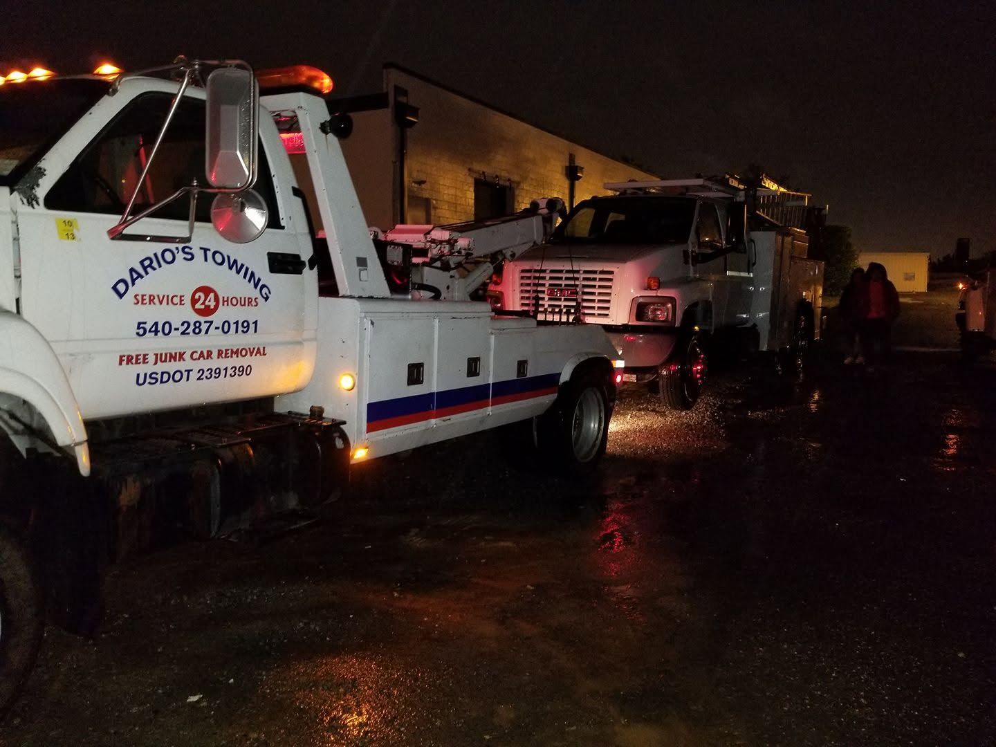 Tow truck towing a truck on a wet road at night, with emergency lights reflecting on the pavement