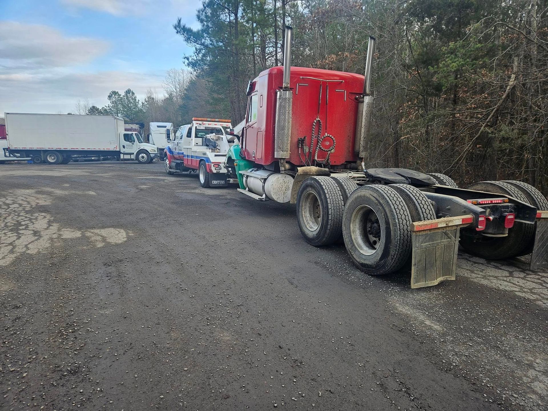 Red semi-truck parked on a gravel lot beside several other trucks and a wooded area