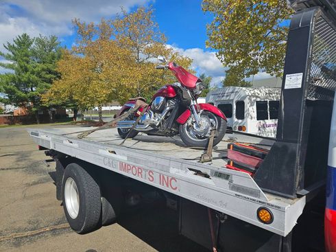 Red motorcycle strapped on a flatbed tow truck outdoors near trees and buildings.