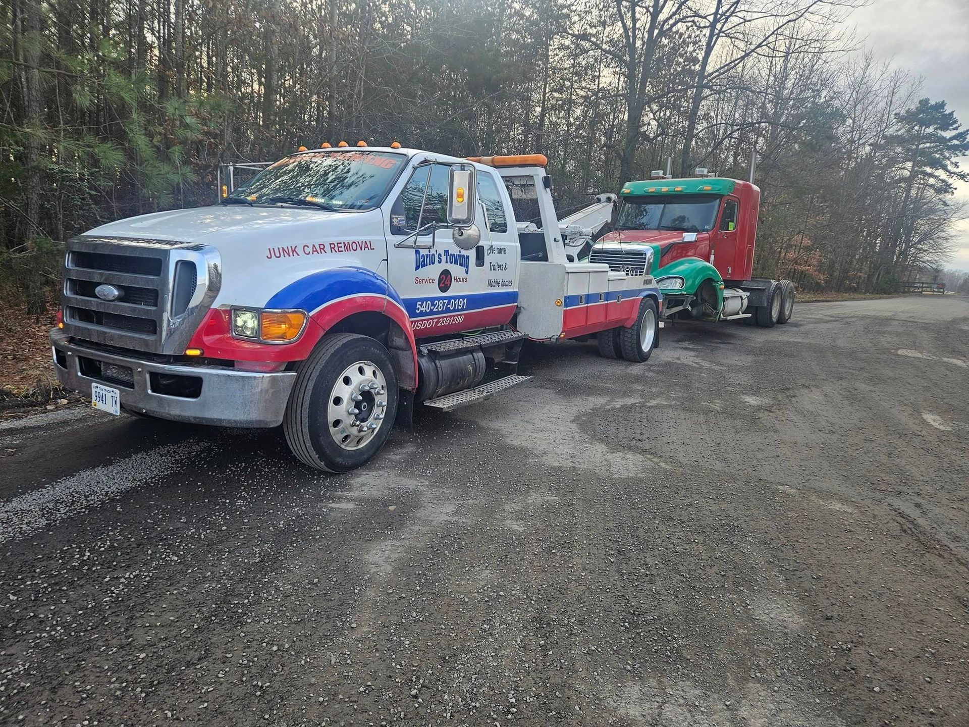 Tow trucks parked on a gravel roadside, one silver with red and blue markings, one green and red.