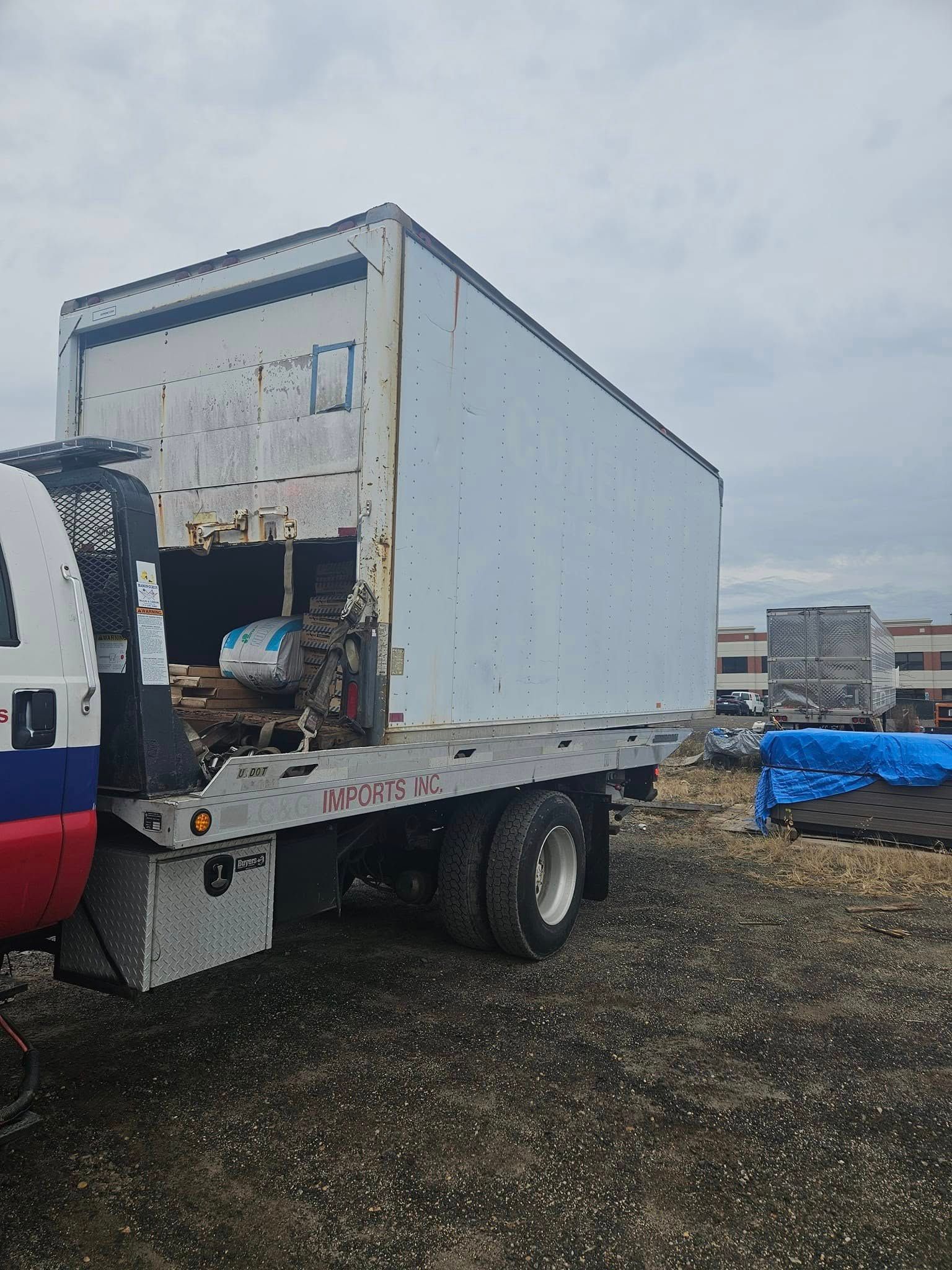 White box truck with rear door open on a gravel lot, with another truck partially visible nearby.
