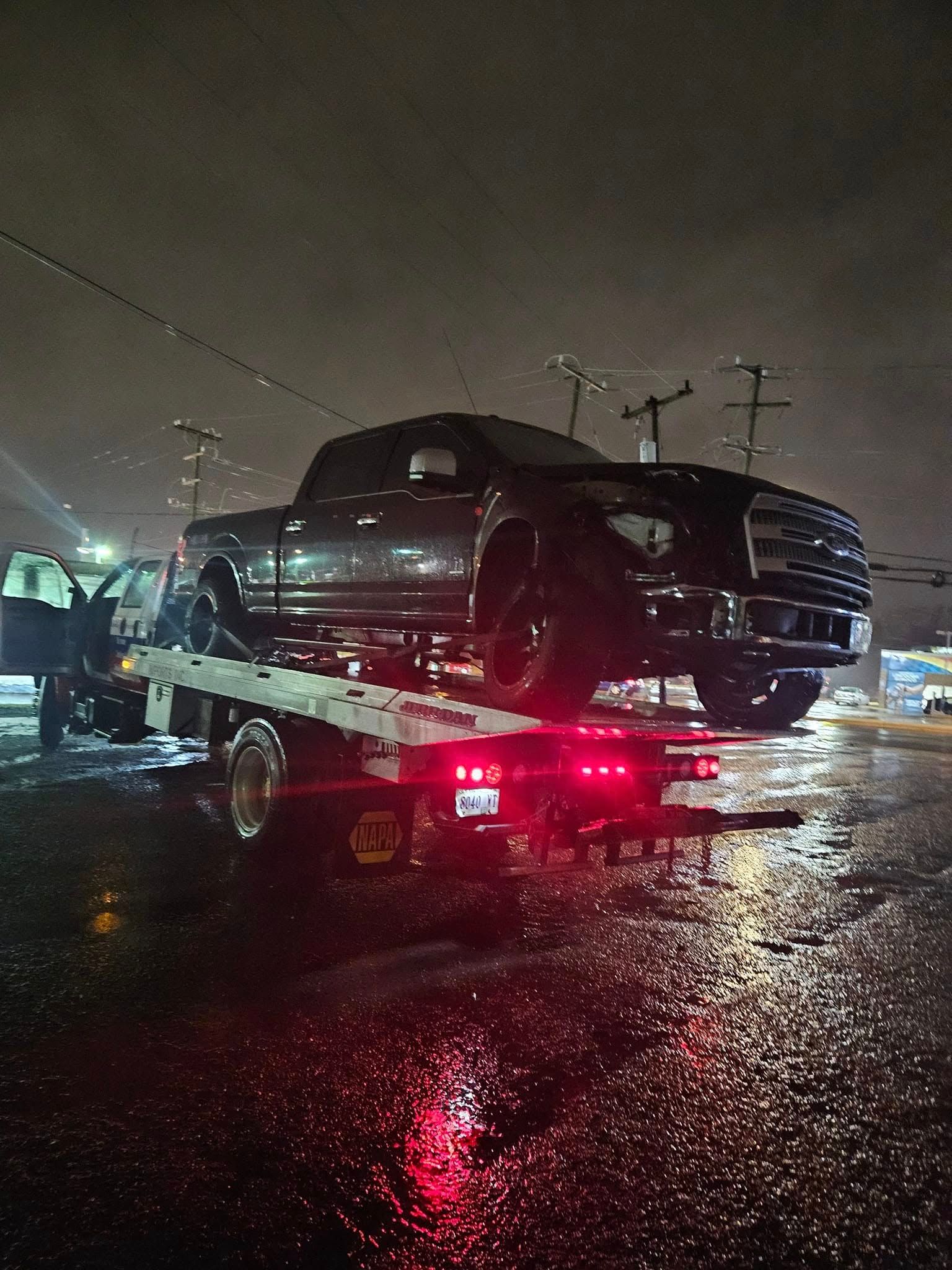 Tow truck hauling a dark SUV on a wet city street at night, with red lights reflecting on the road