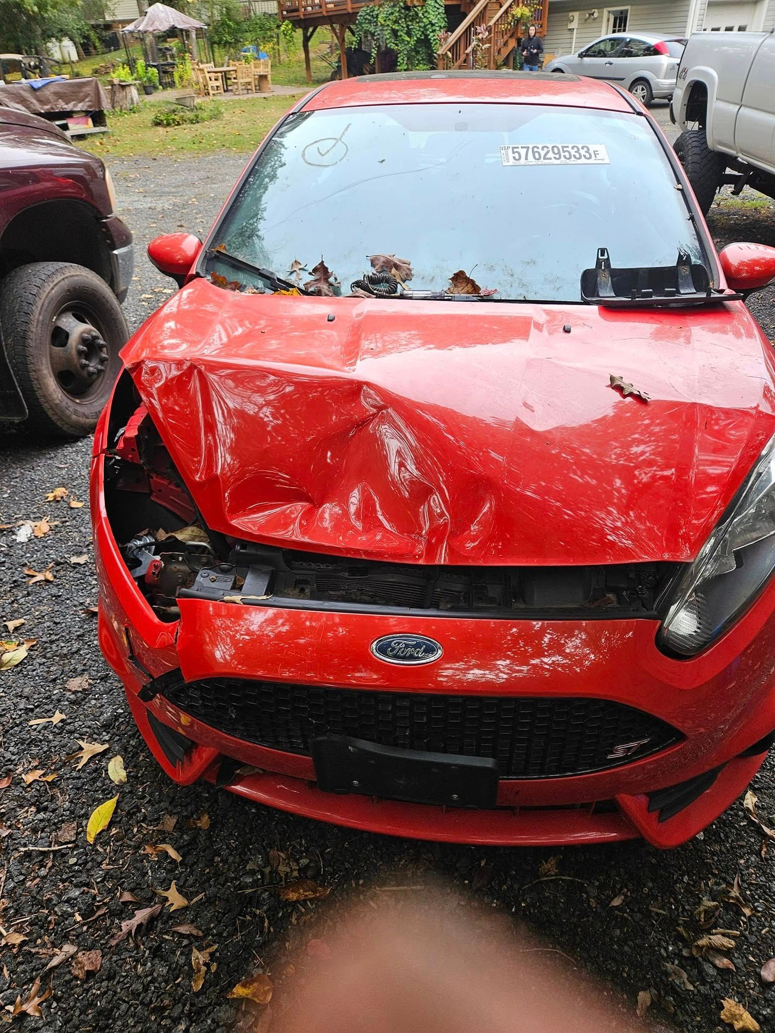 Red compact car with a crumpled hood and front bumper, parked on wet pavement after damage