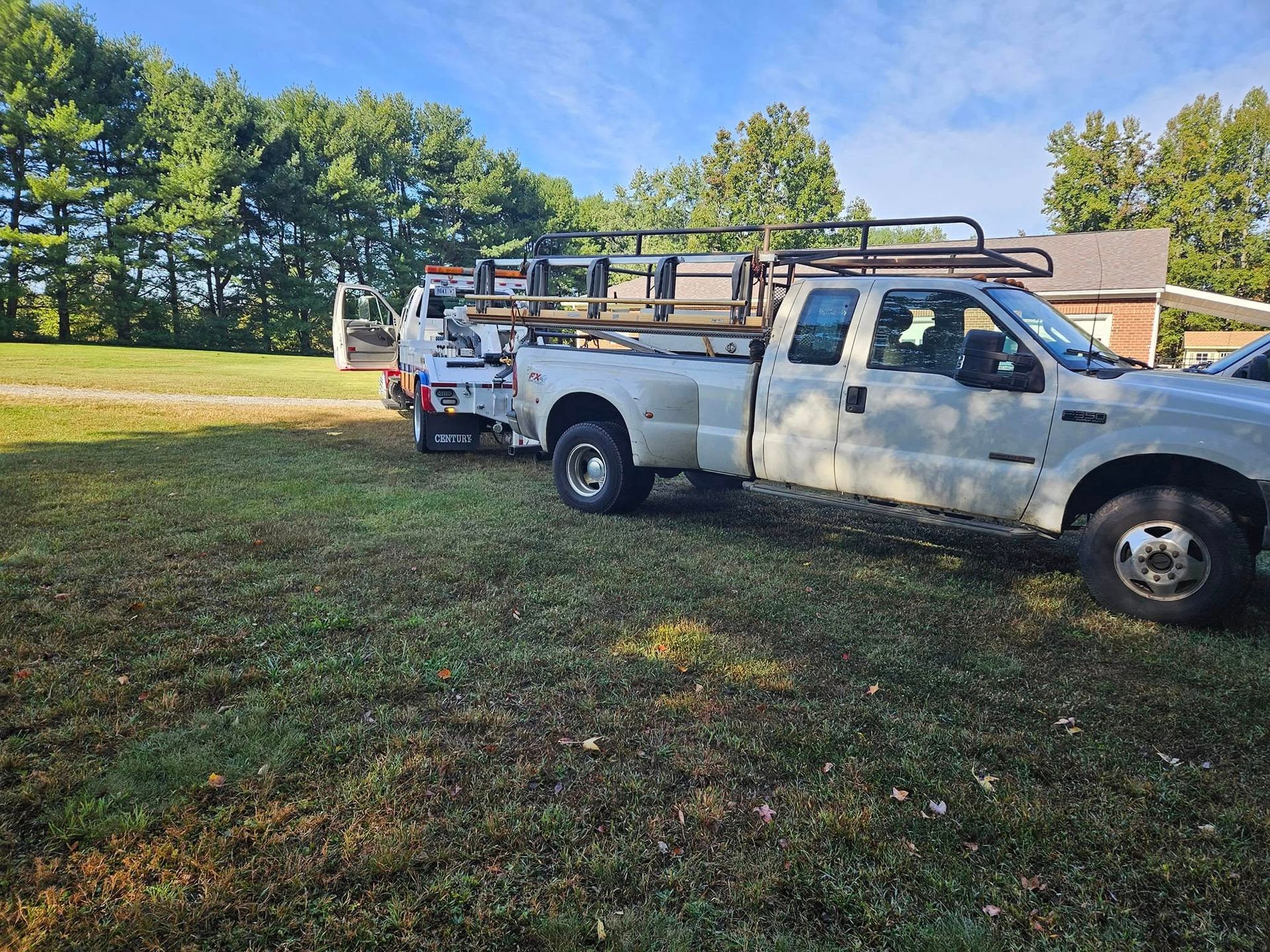 Two white utility trucks parked on grass beside trees and a house under a blue sky