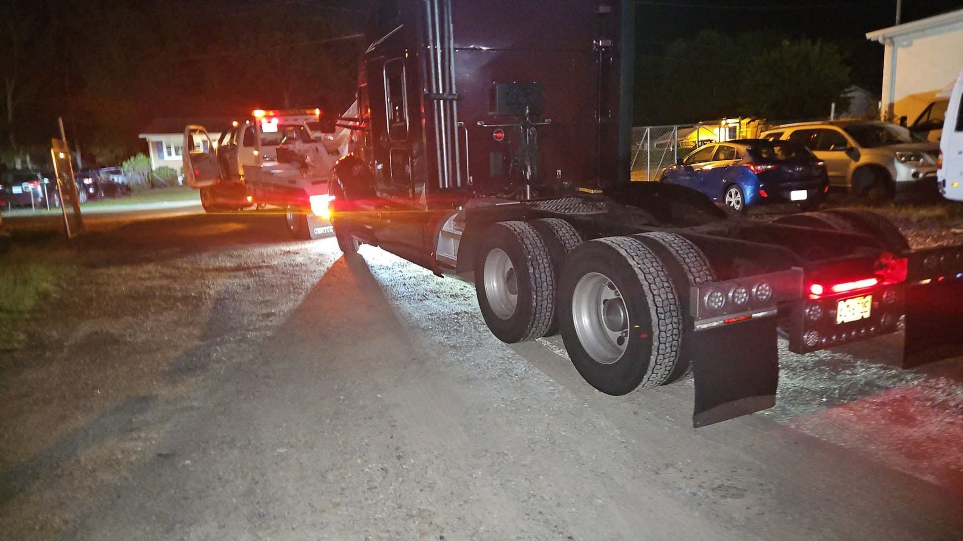 Two semi-trailers at night on a snowy road, with red taillights and emergency vehicles in the distance.