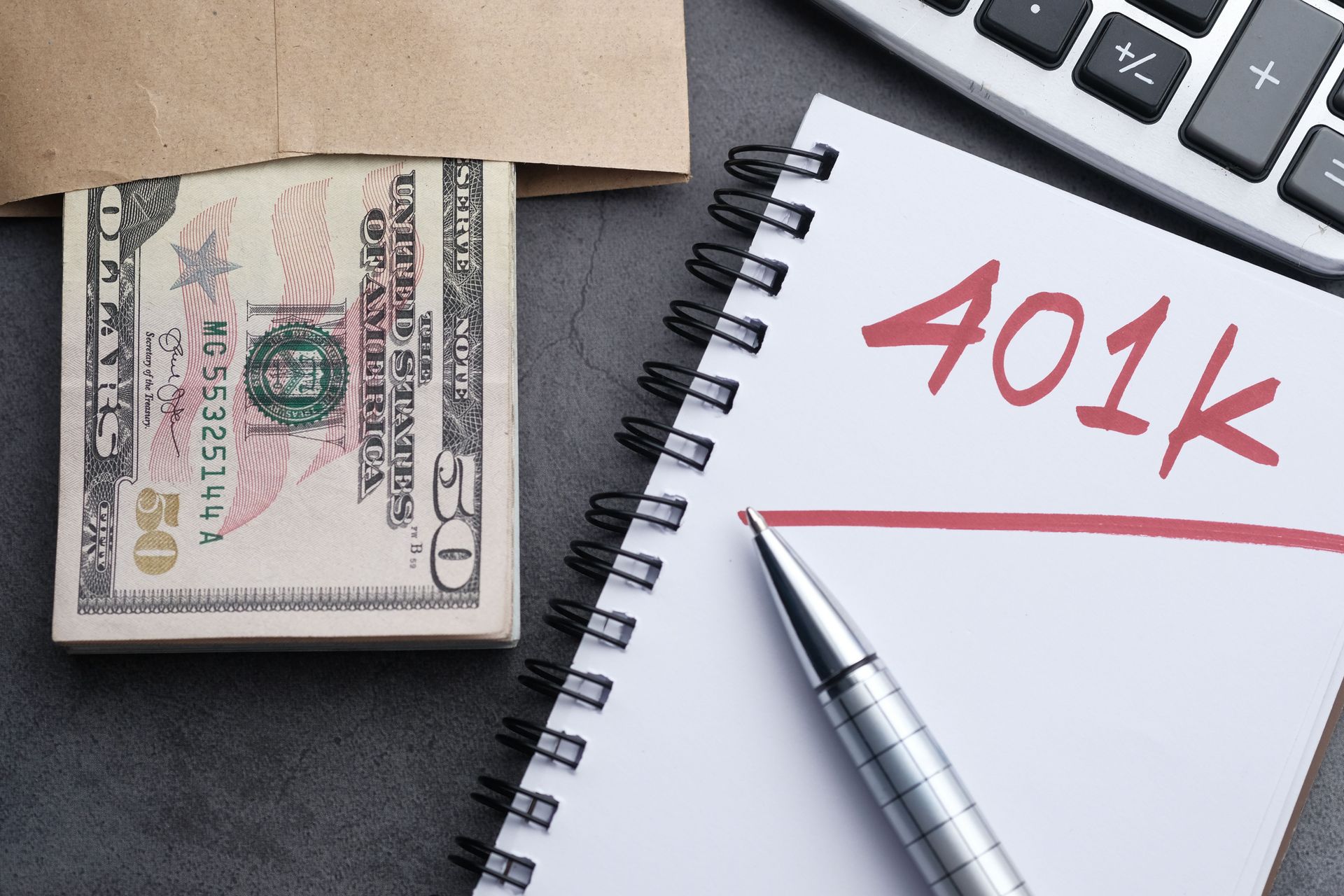 Stack of cash beside a notebook labeled “401k” with a pen on a desk.