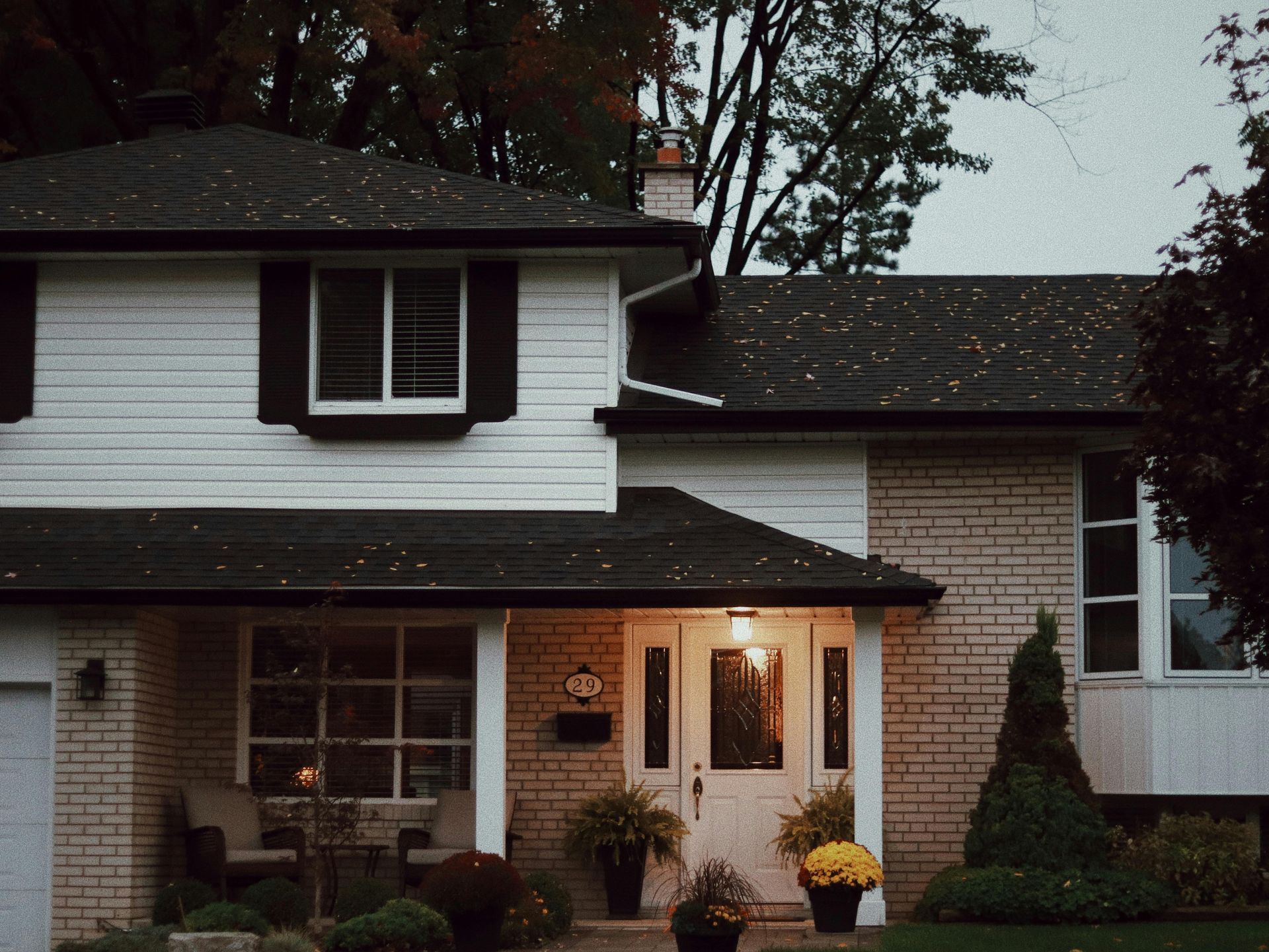 Suburban house at dusk with lit porch lights, white siding, brick facade, and dark roof.