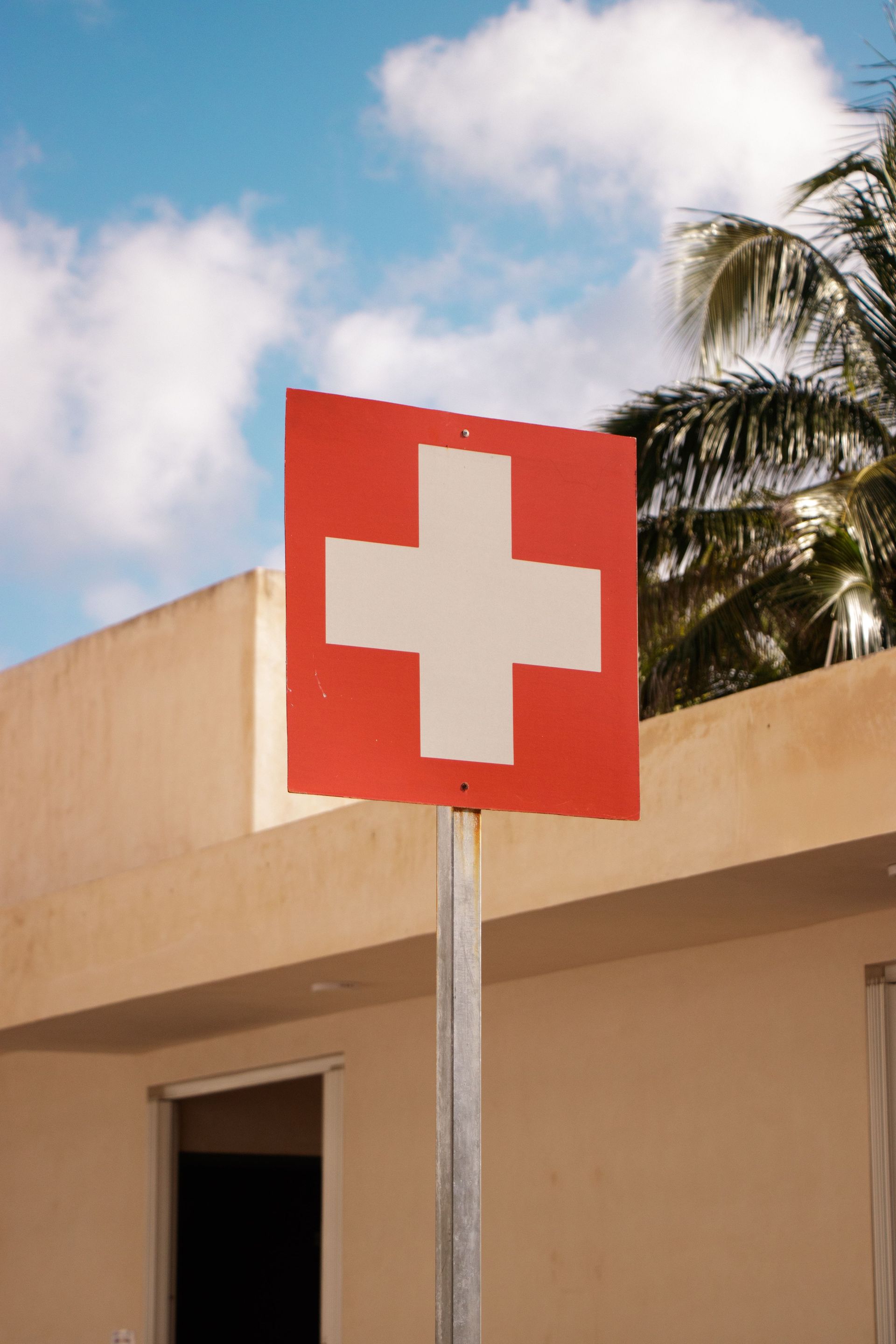 Red sign with a white cross on a pole outside a beige building under a blue sky.