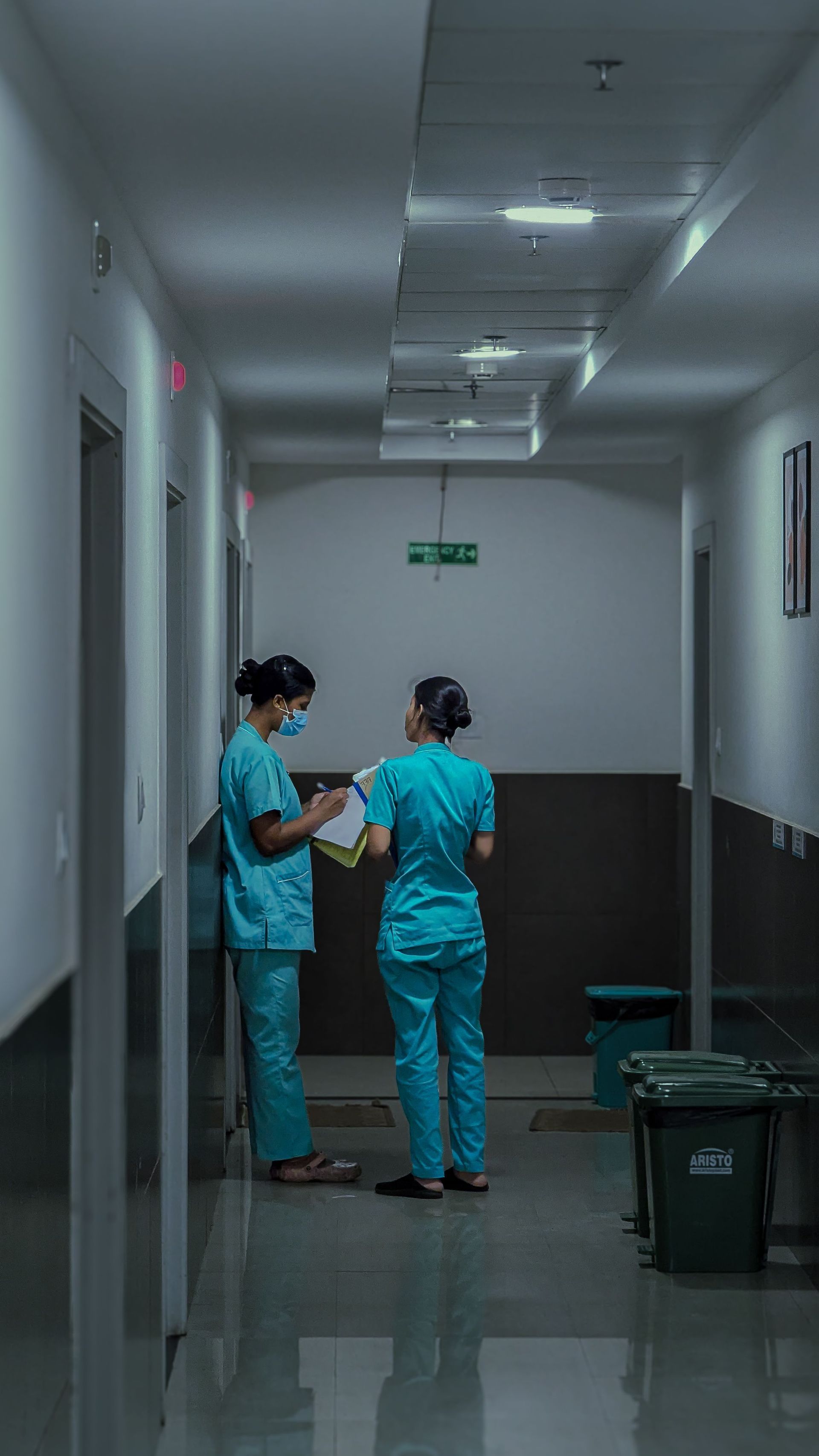 Two nurses in blue scrubs stand in a hospital corridor, talking near a doorway.