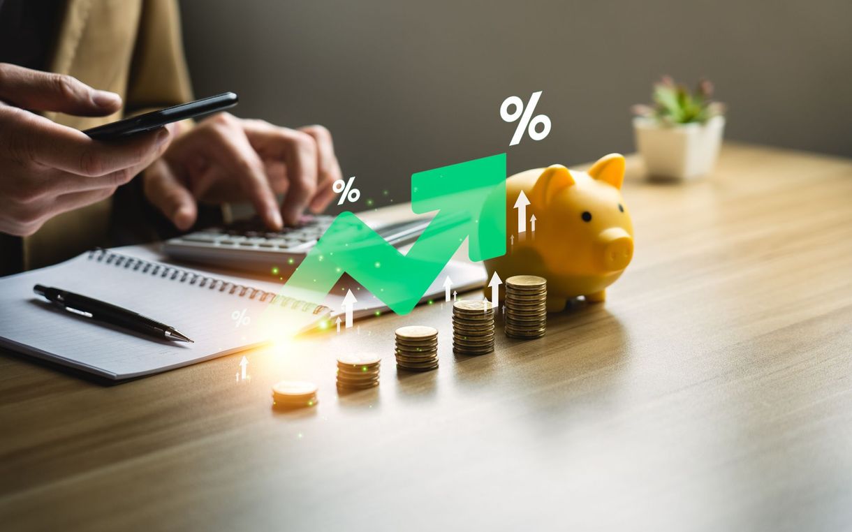 Hand using phone beside rising coins, green arrow chart, and yellow piggy bank on desk