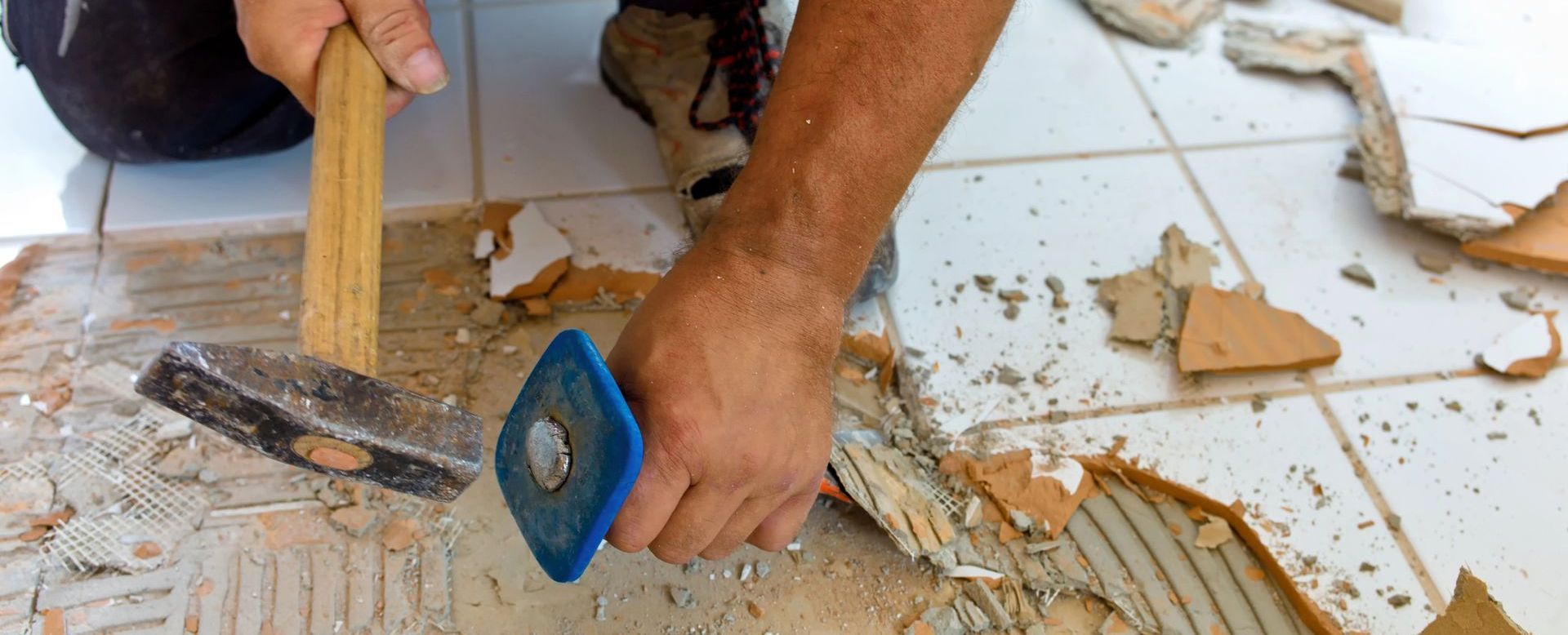 A man is using a hammer and a spatula to remove tiles from the floor.