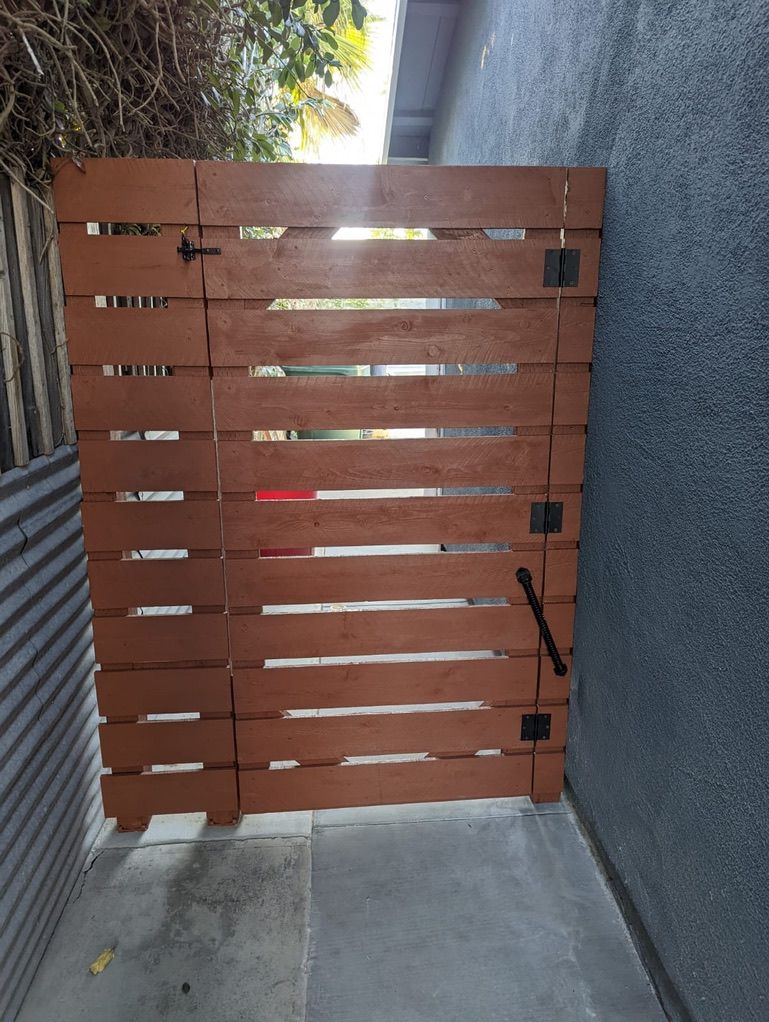 Wooden gate painted brown with black hardware, next to a gray stucco wall and a corrugated metal fence.
