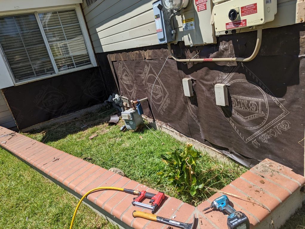 Construction site: brown siding, foundation, brick border, lawn. Tools in foreground.