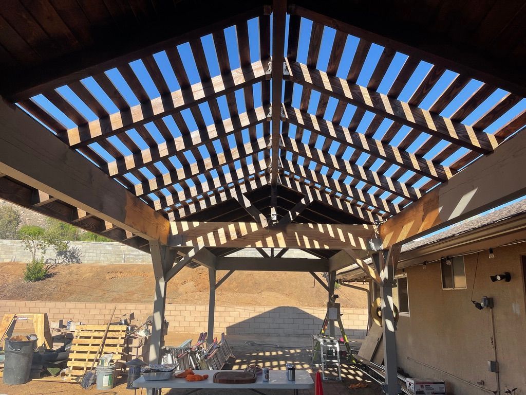 Wooden pergola roof casting shadows on a backyard patio, with construction materials visible below.