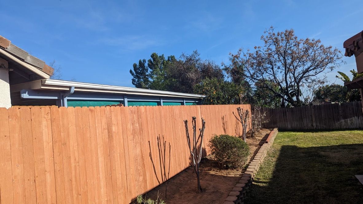 Wooden fence, yard, and blue sky. A house is visible with a detached garage-like structure and some greenery.