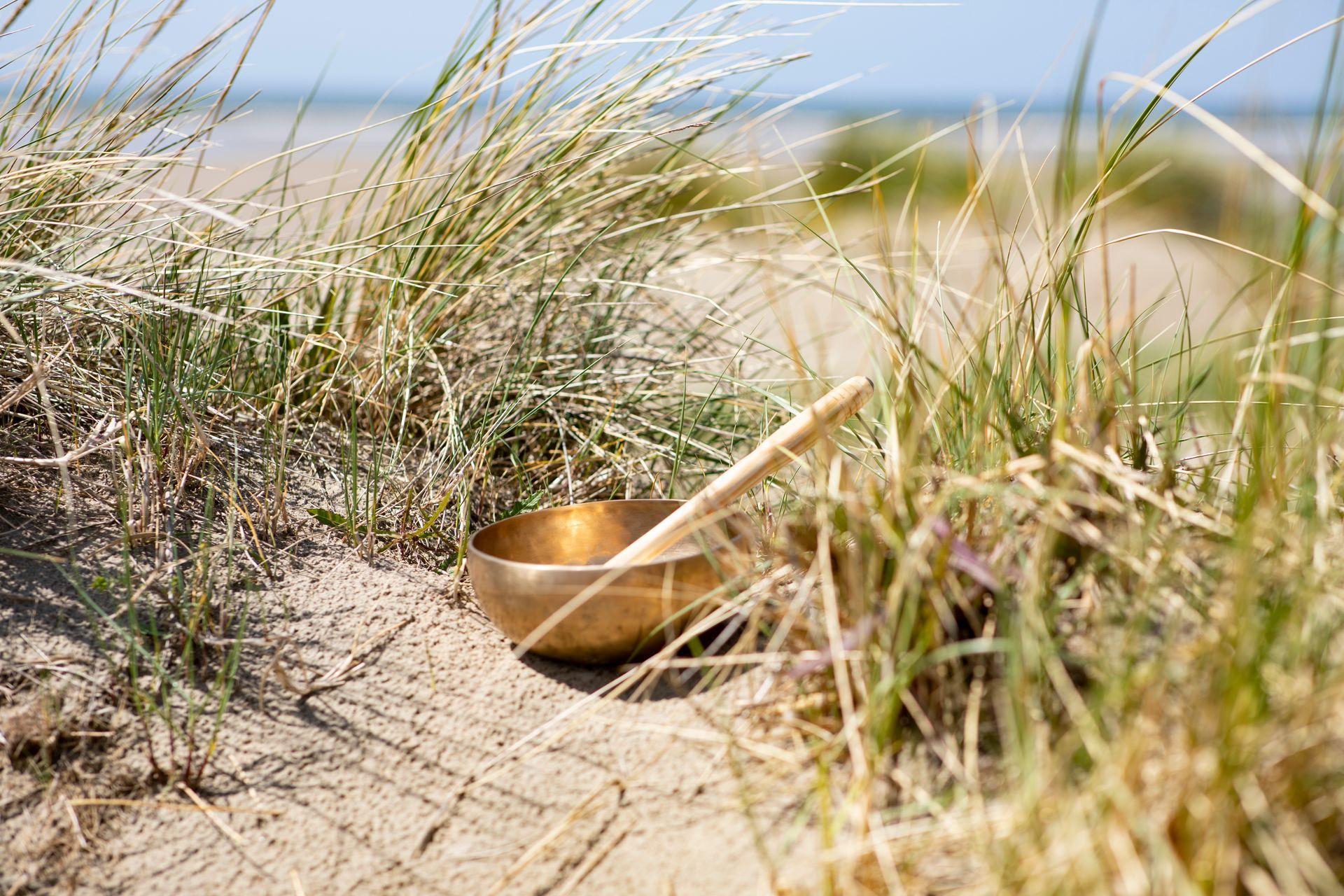 Een koperen klankschaal met een houten klopper staat in het zand op een strand
