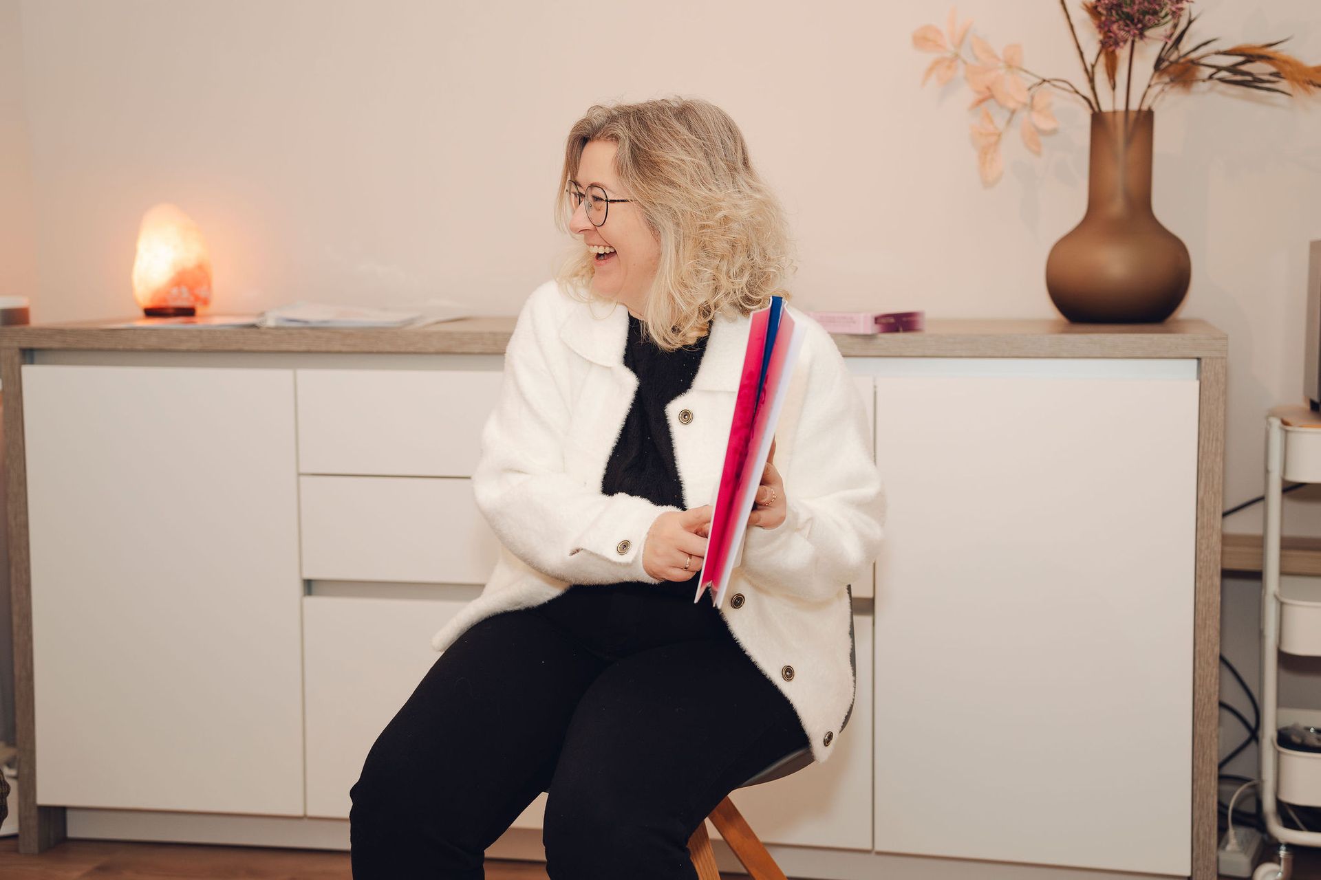 Een vrouw zit op een stoel in een kamer en houdt een roze voorwerp vast.