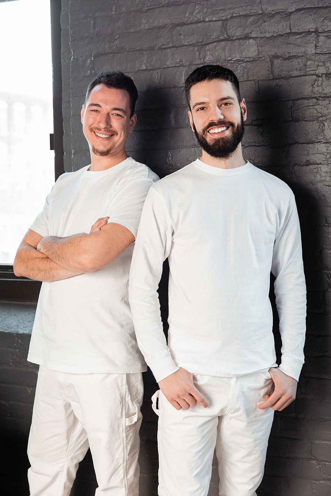 Two men in white outfits smiling against a black brick wall