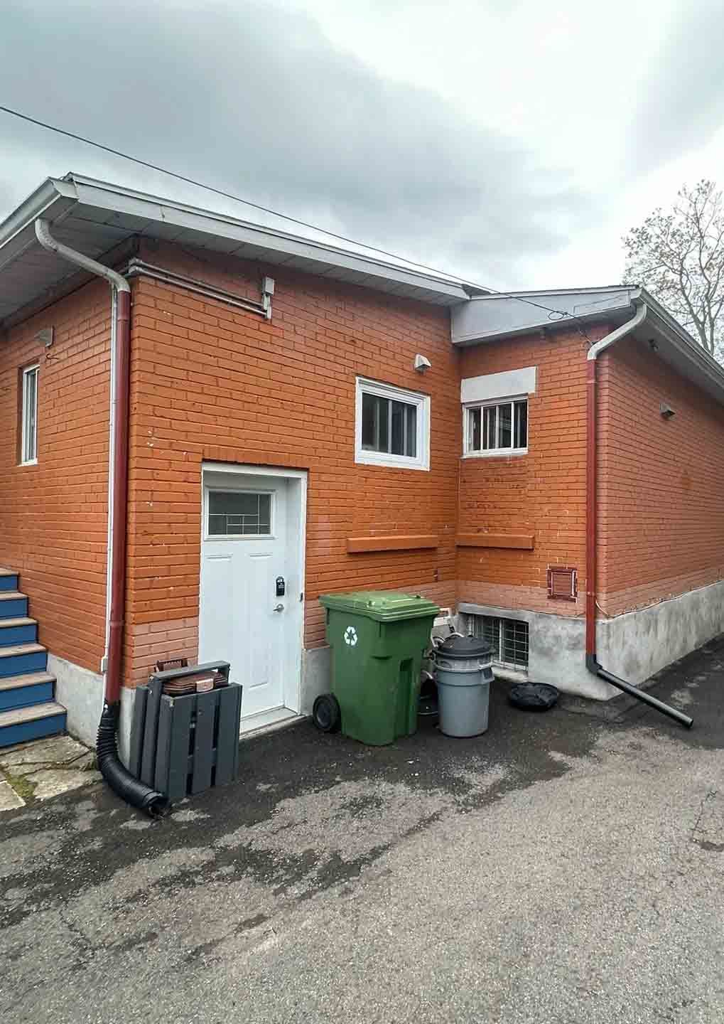 Brick building exterior with white door, windows, green bins, and blue steps under an overcast sky