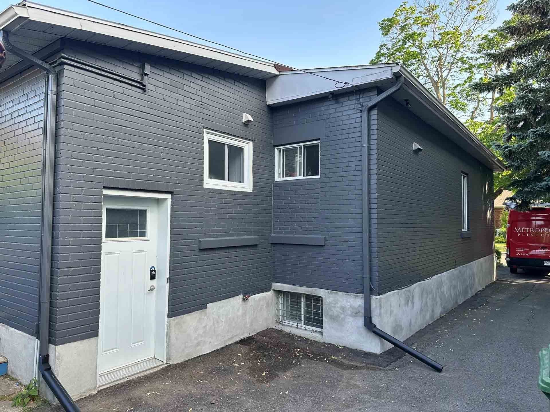 Dark gray brick building with a white door and windows beside a narrow driveway