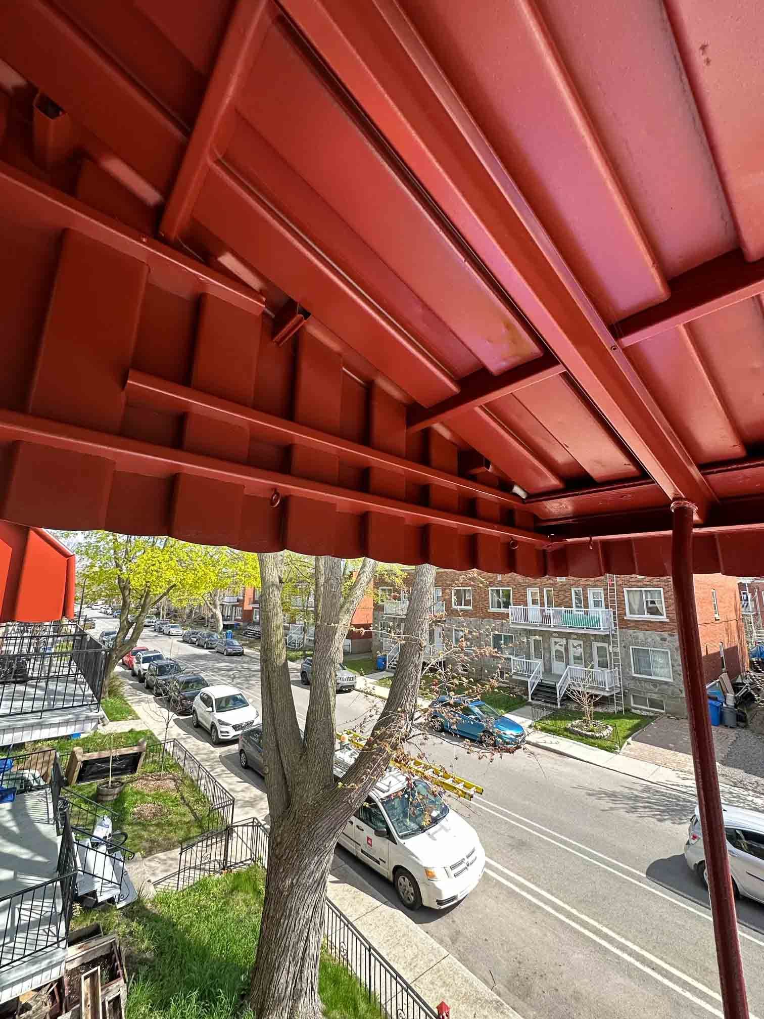 Red metal roof over a street with parked cars, trees, and buildings below