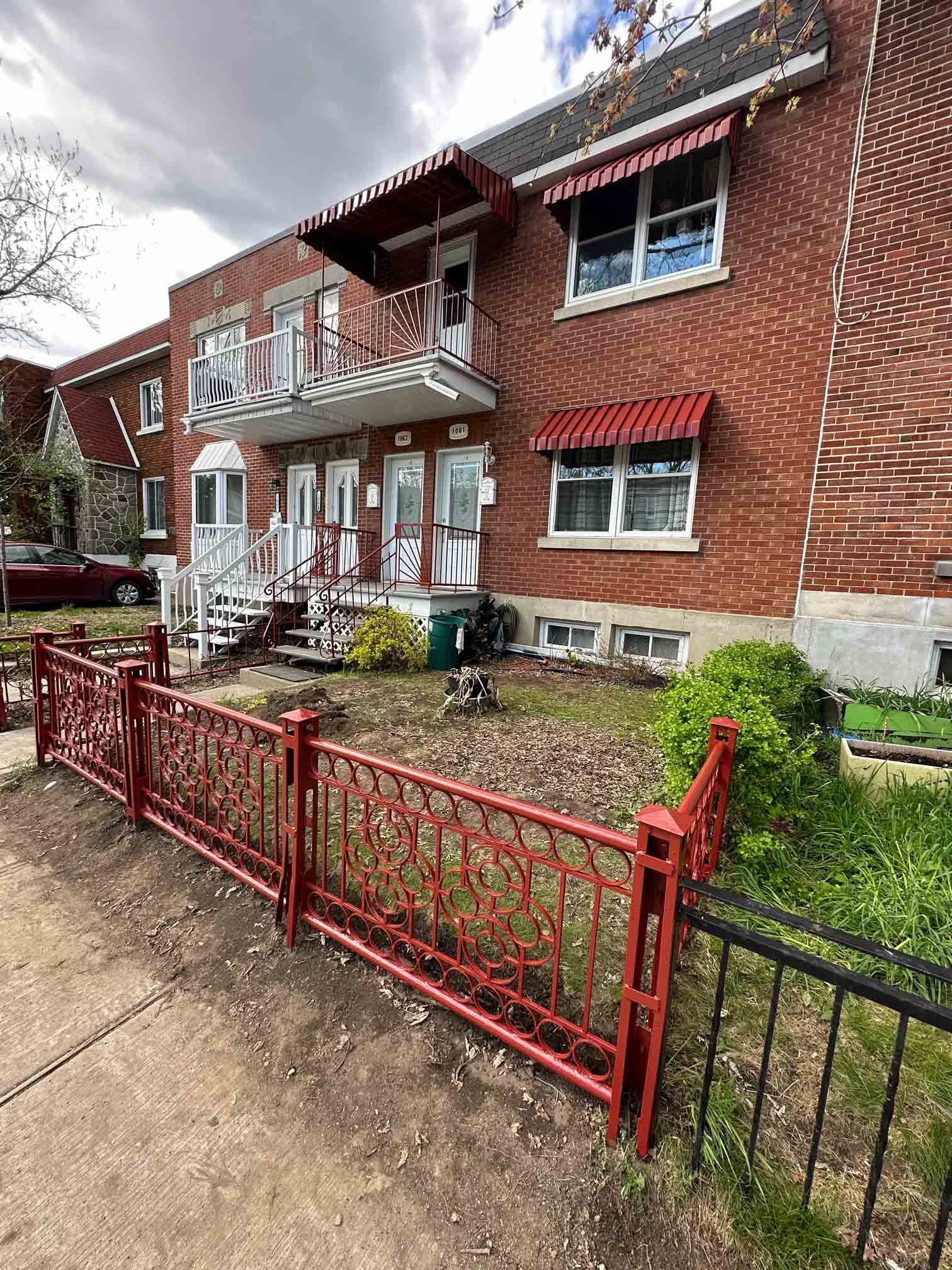 Brick apartment building with red awnings, small front porch, and red metal fence by a walkway