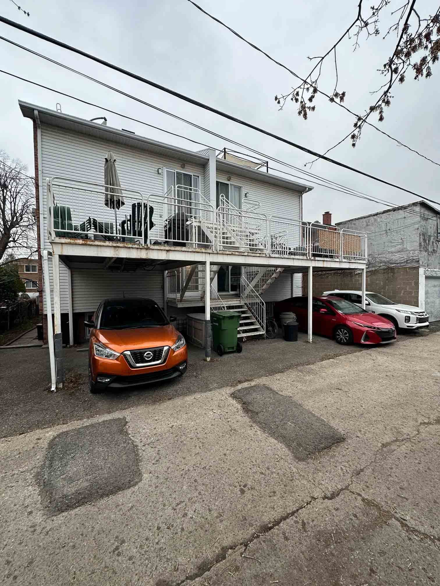 Two-story apartment building with cars parked under covered spaces and a red car beside it