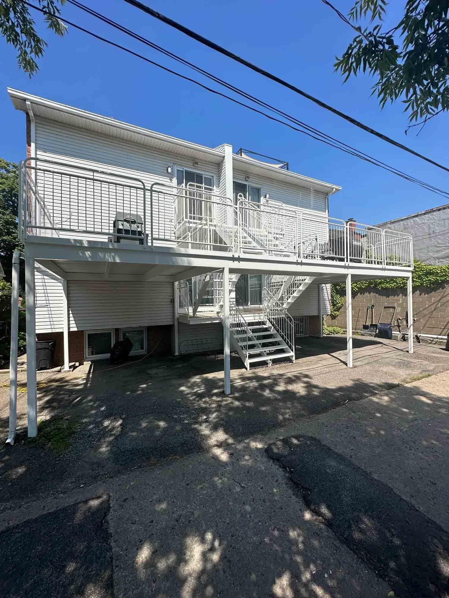 Two-story white house on stilts with an exterior staircase and carport beneath, under a clear blue sky.