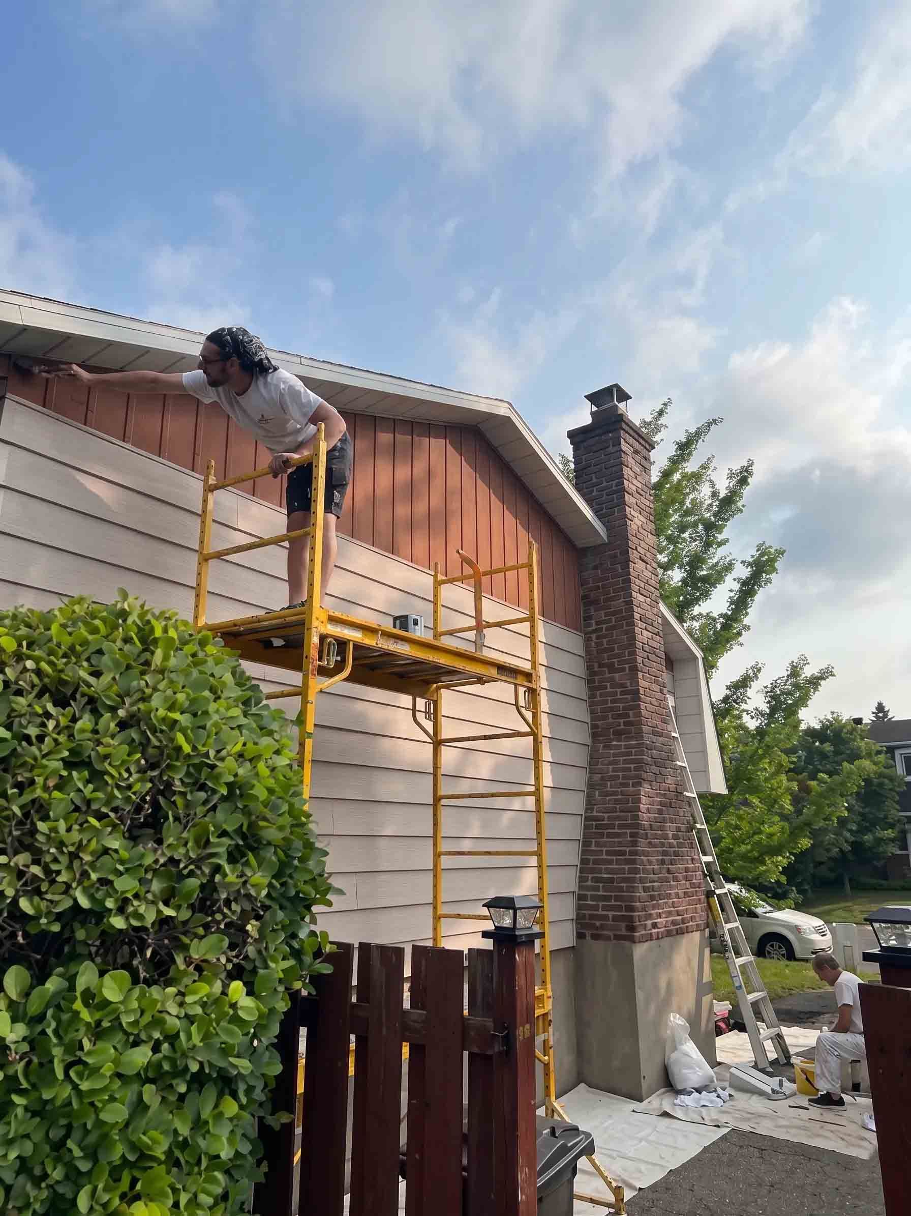 Worker on a yellow scaffold painting a wooden house exterior beside a brick chimney.