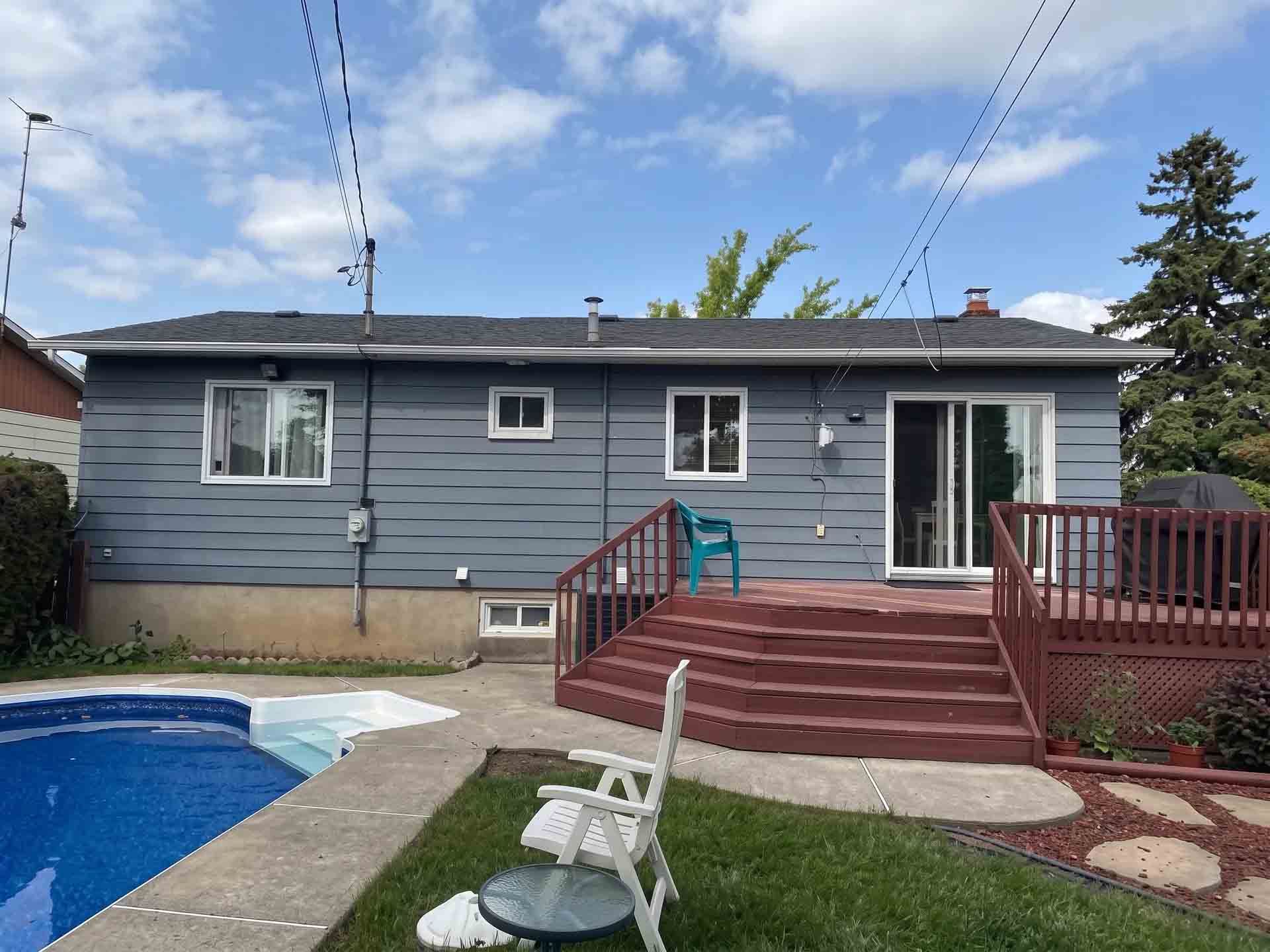 Backyard with blue pool, white chair, red deck, and gray house under a bright sky