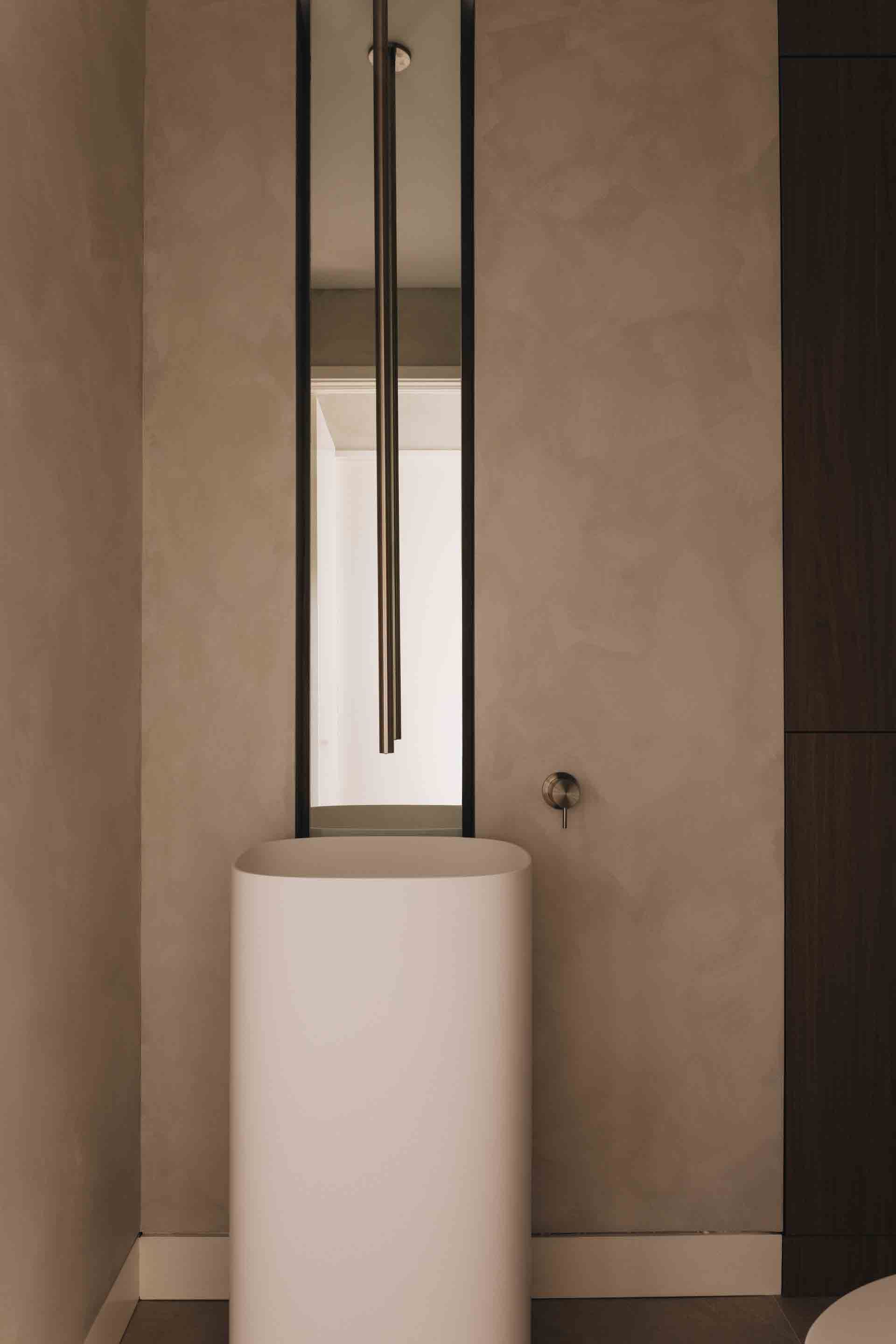 Minimalist bathroom with a white pedestal sink beneath a tall mirror and dark vertical fixture