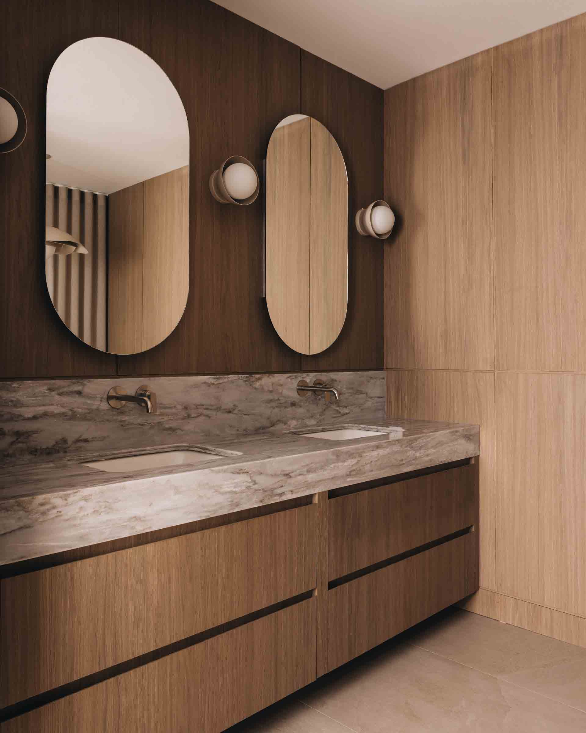 Modern bathroom vanity with wood paneling, marble countertop, and oval mirrors with wall sconces.