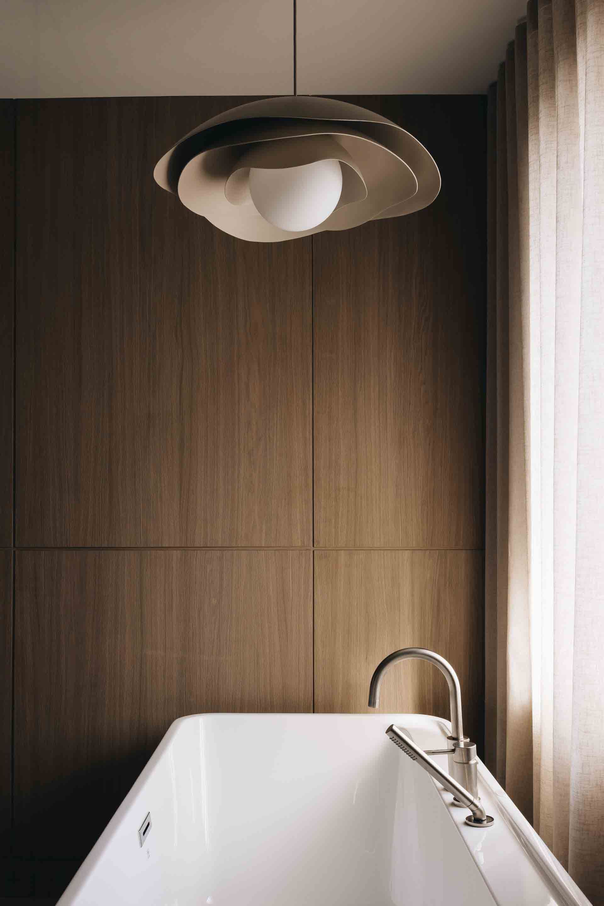 Modern bathroom with a freestanding tub, wood-paneled wall, ceiling light, and sheer curtain.