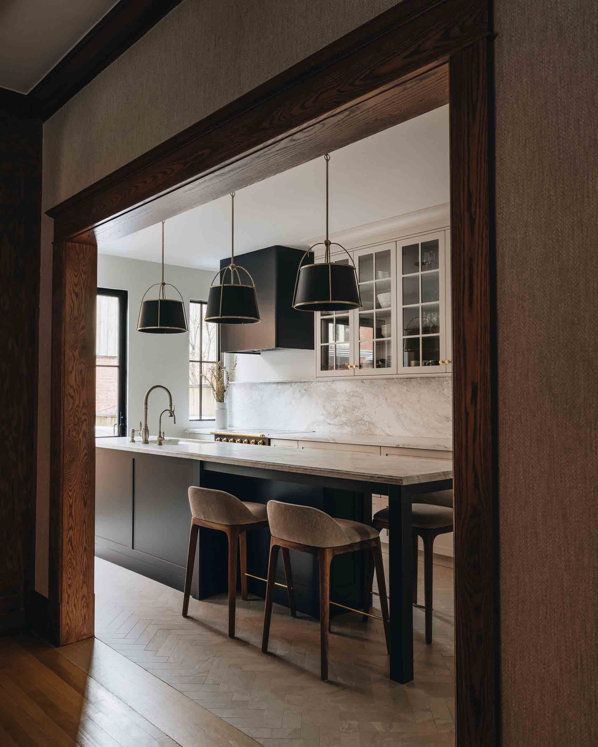 Open kitchen viewed through a doorway, with island stools, hanging lights, and white cabinets.