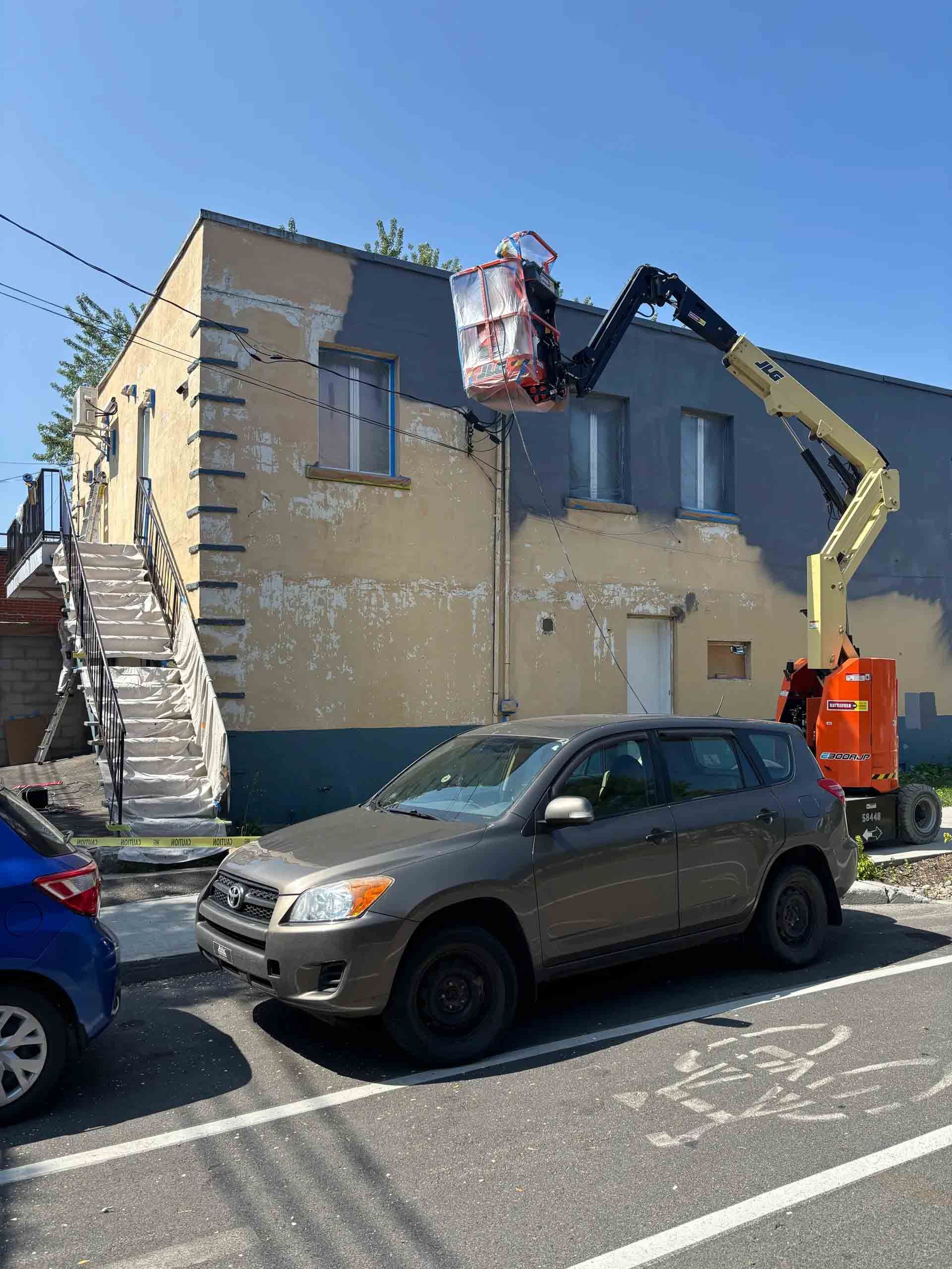 Un ouvrier, à bord d'une nacelle élévatrice, retire une enseigne endommagée d'un immeuble beige dans une rue ensoleillée, avec des voitures garées en contrebas.