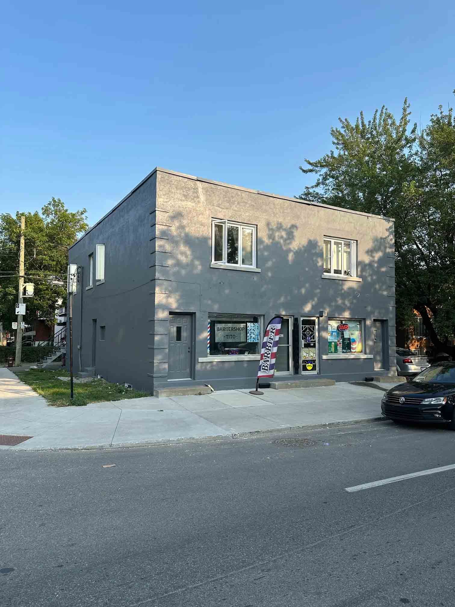 Gray two-story corner building with storefront windows, a sidewalk sign, and a parked car on a quiet street.