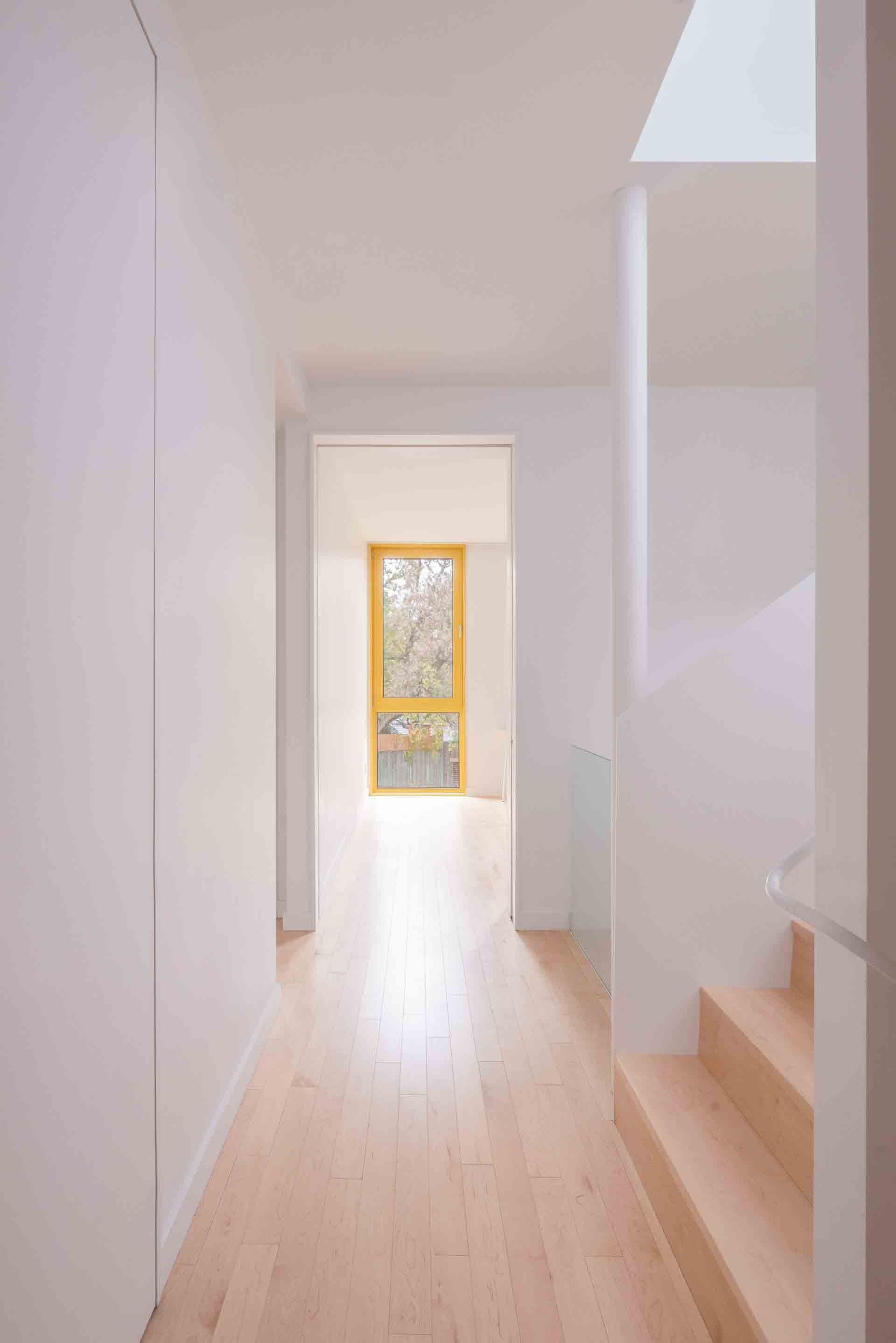 Couloir minimaliste et lumineux avec parquet clair, murs blancs, escalier et une porte baignée de soleil au fond