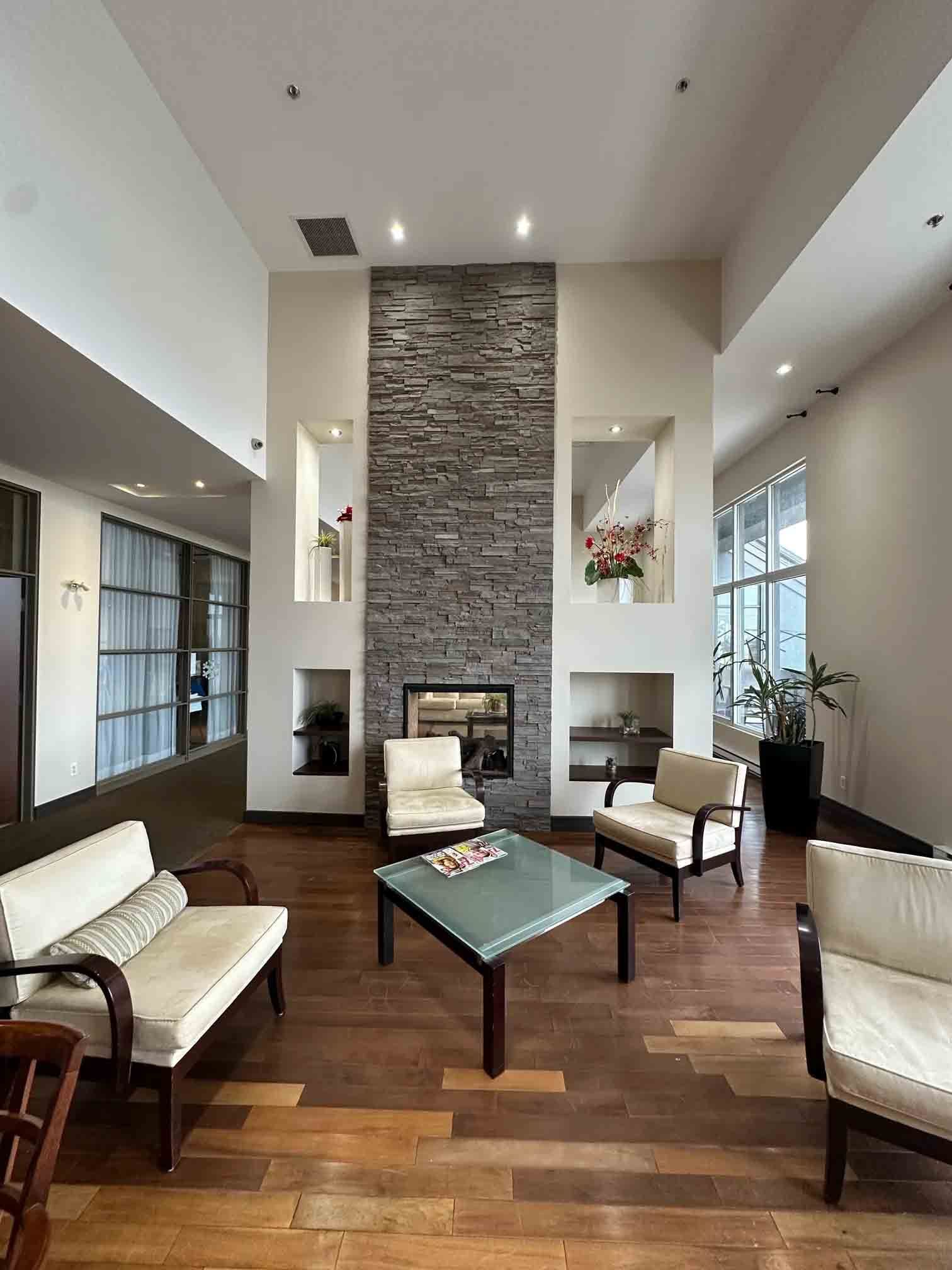 Modern living room with stone fireplace, high ceiling, and beige armchairs around a coffee table.