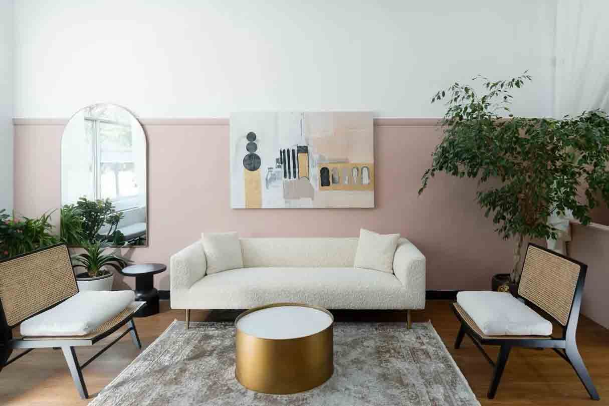 Modern living room with cream sofa, gold coffee table, two chairs, and large green plant against a pink wall