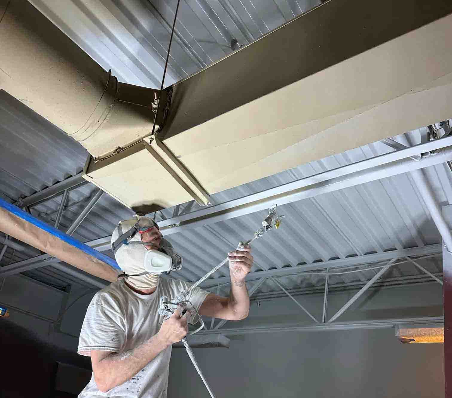 Worker in mask spraying paint on ceiling beams inside an industrial building