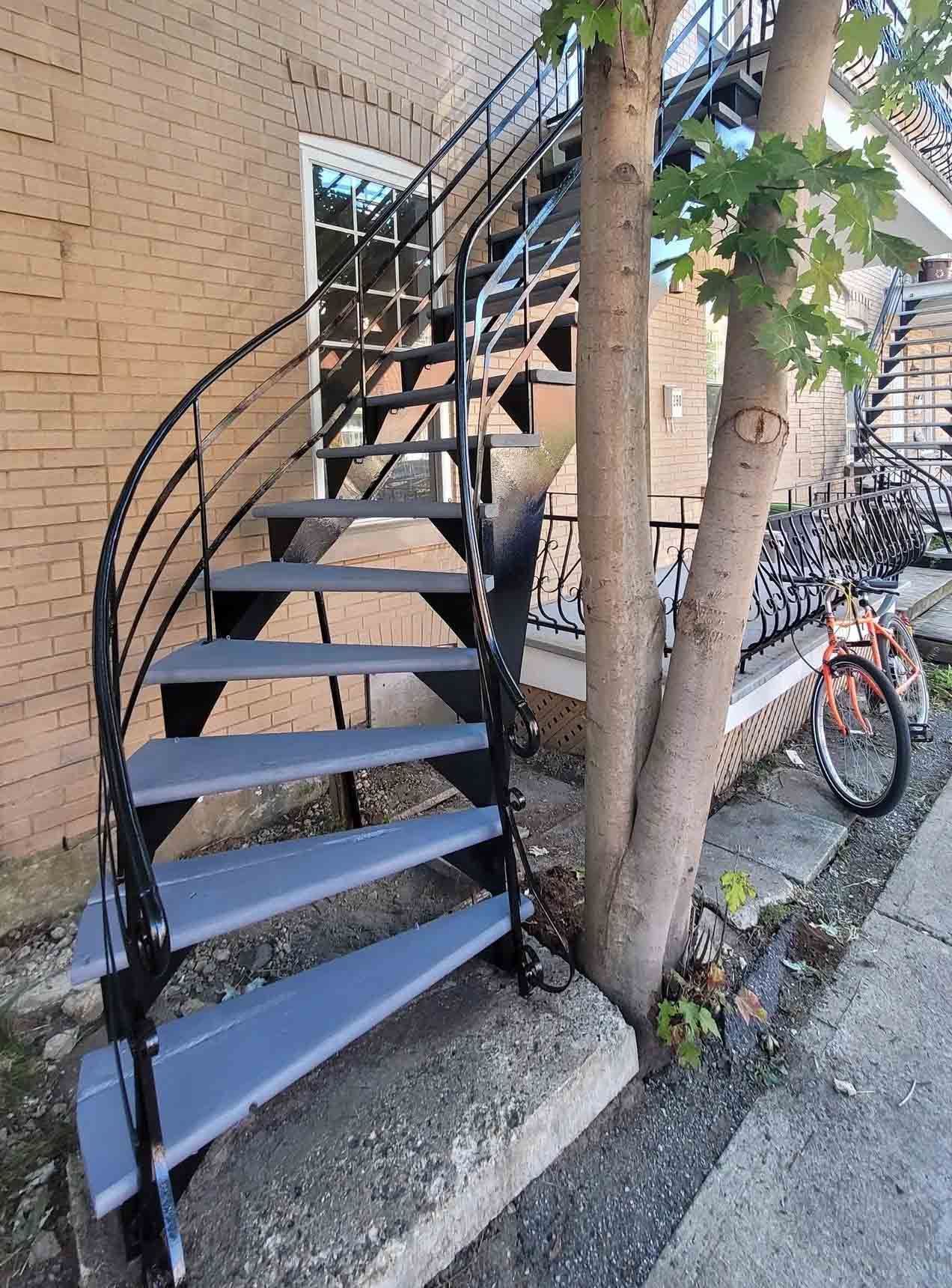 Black metal spiral staircase beside a brick building, with trees and a bicycle nearby.